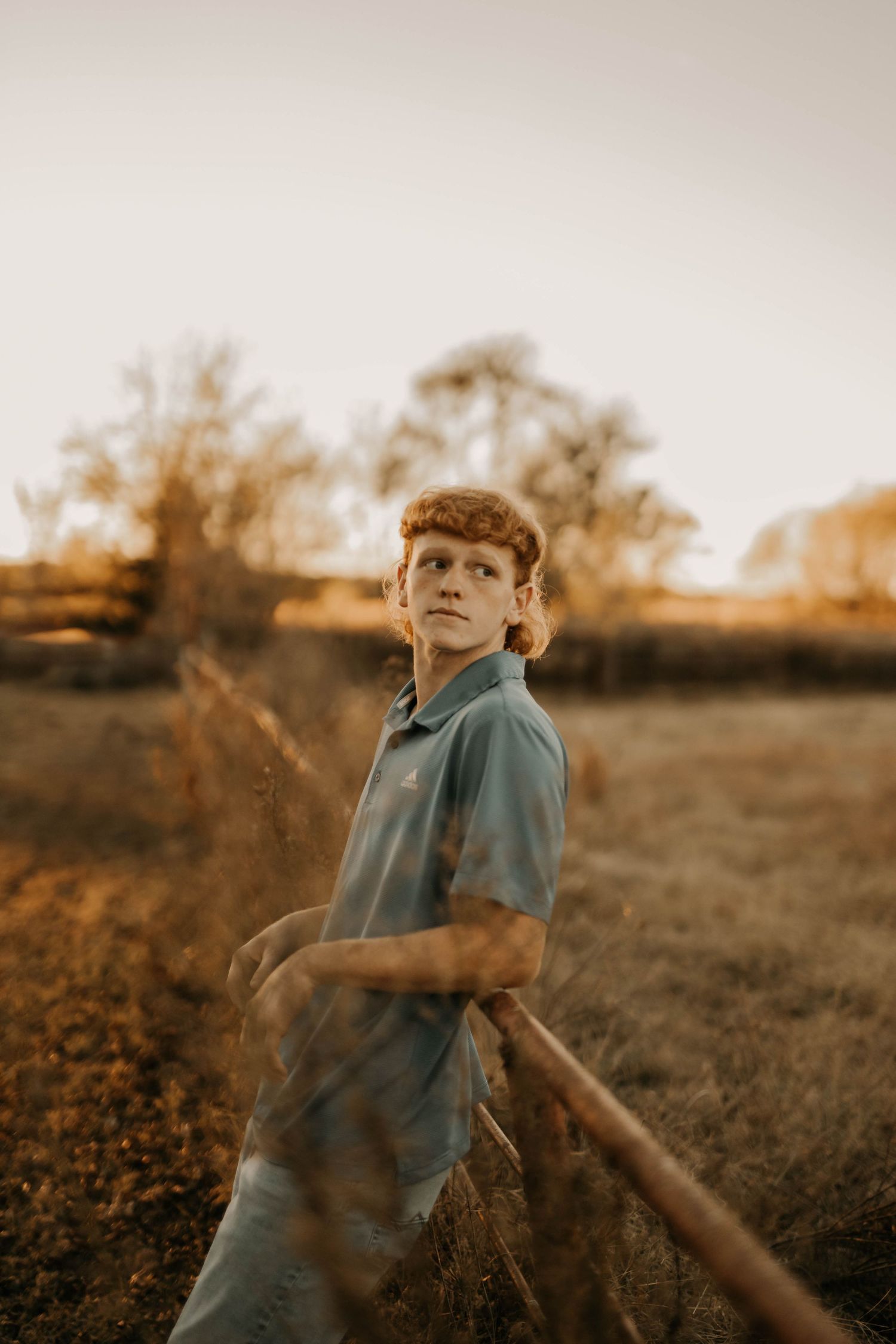 Person in blue polo leans on wooden fence during golden hour in rural setting.