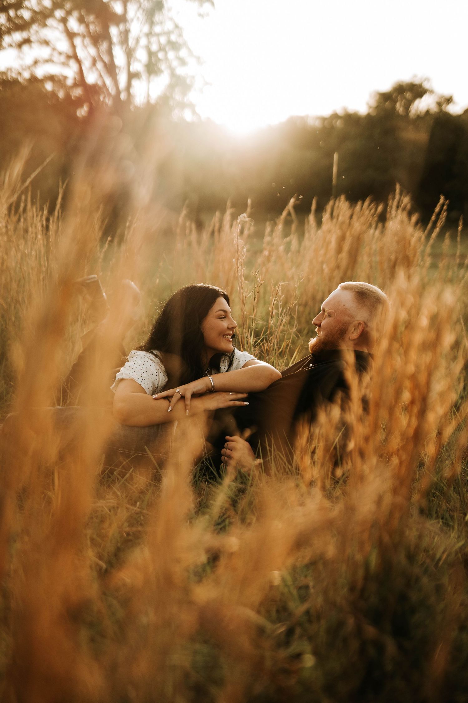 Couple laughing together while sitting in golden tall grass during sunset.