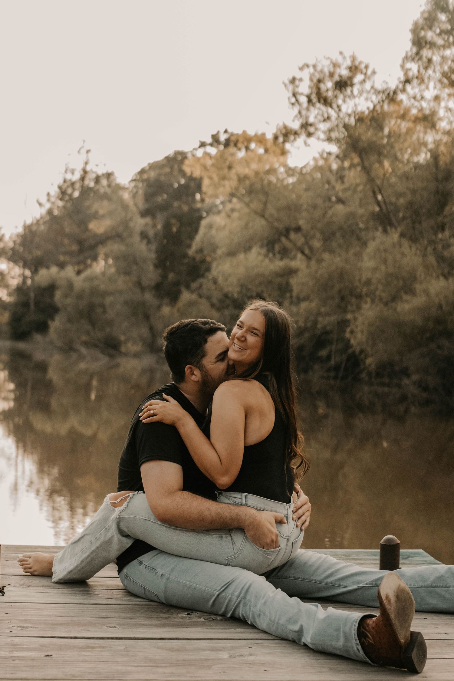 Couple cuddling intimately on a wooden pier with a scenic lake and trees in the background.
