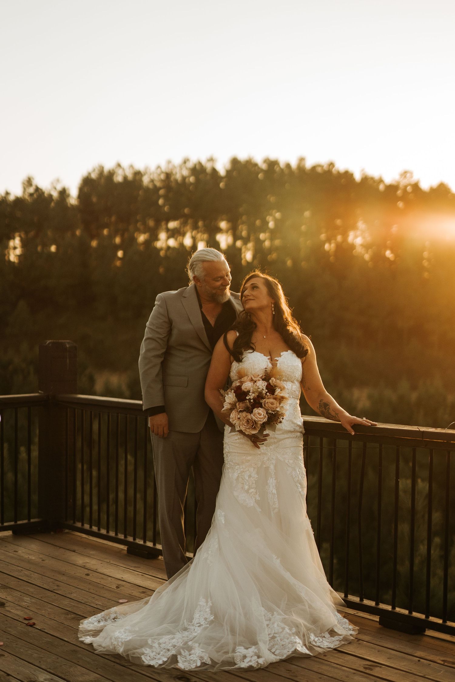 Bride and groom pose on wooden deck at golden sunset with forest backdrop, bride holding bouquet in lace wedding gown.