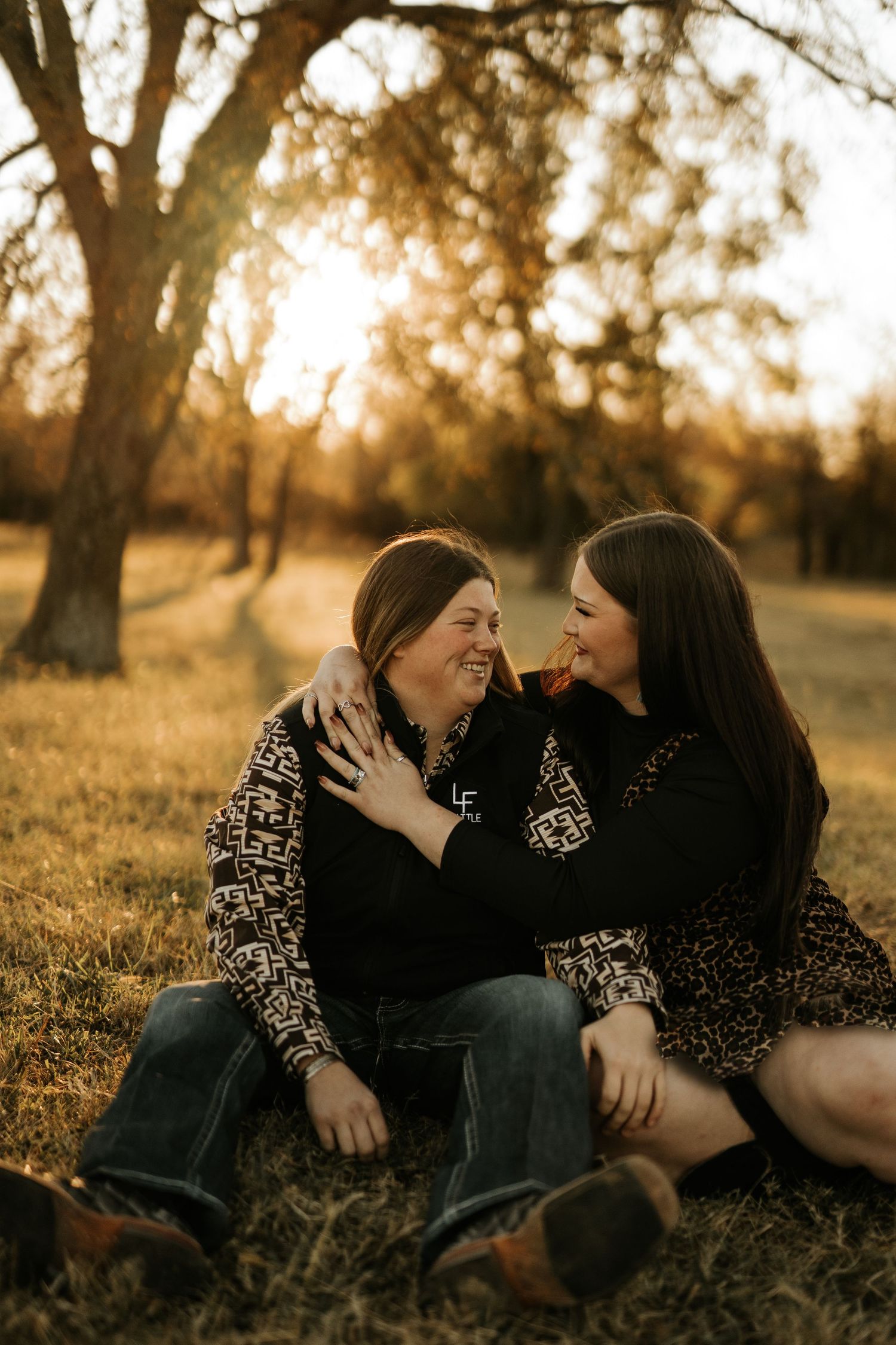 Two women sitting on grass at golden hour, smiling and embracing in a sunlit park with autumn trees.
