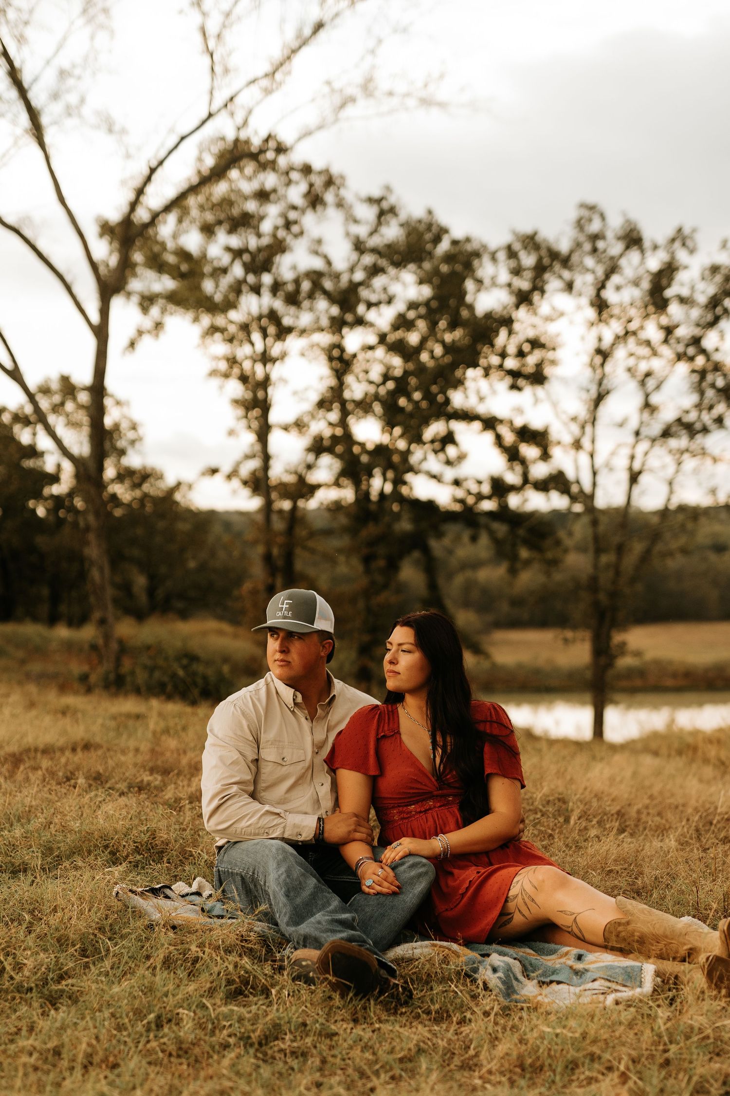 Couple sitting on a blanket in a grassy field, man in cap and woman in red dress, with trees and pond in background.