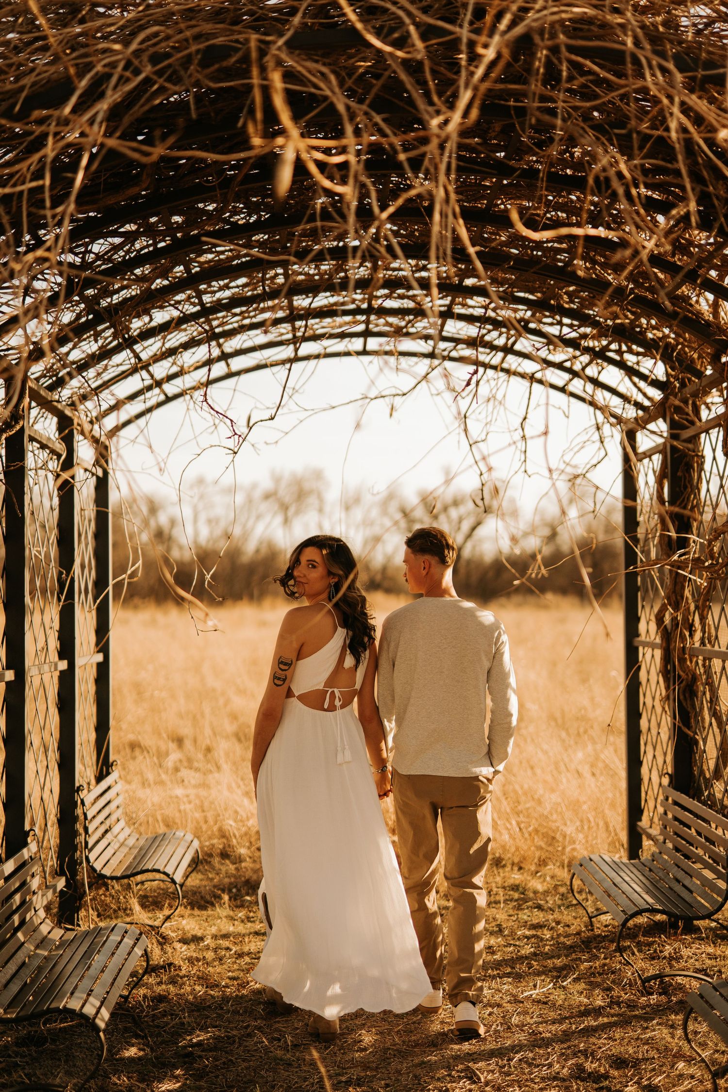 Couple in wedding attire walking under a rustic vine archway in a golden sunlit field.