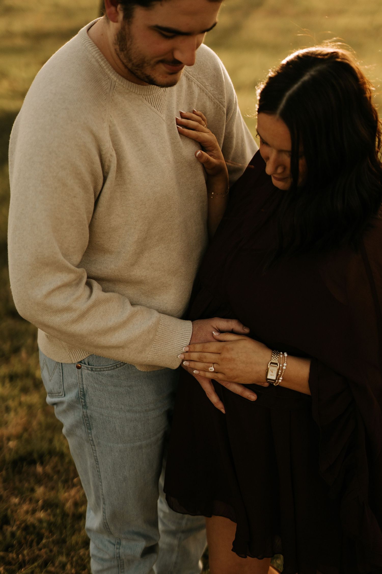 A couple tenderly cradles a pregnant belly outdoors during golden hour, wearing casual attire in a maternity photo session.