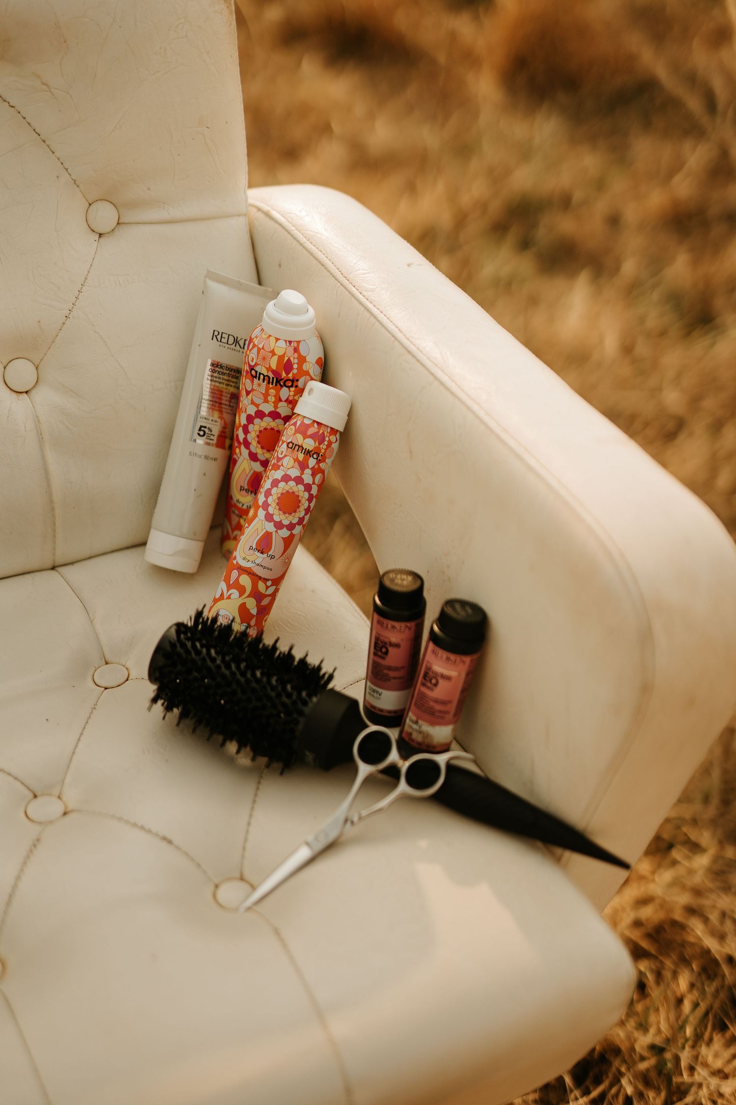 Hair styling tools including a round brush, scissors, and Redken products rest on a tufted white leather chair outdoors.