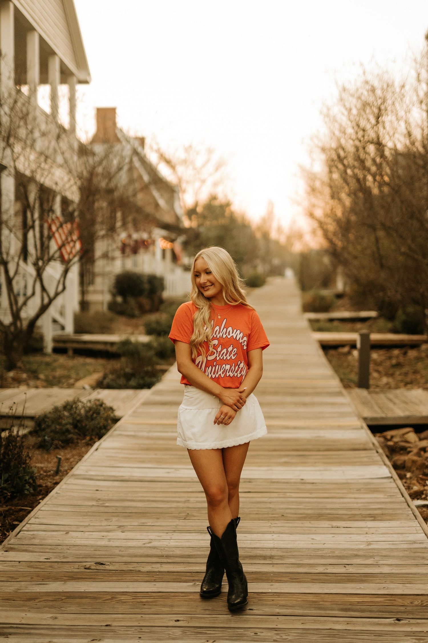 Blonde woman in Oklahoma State University orange tee, white mini skirt, and black cowboy boots on a wooden boardwalk.