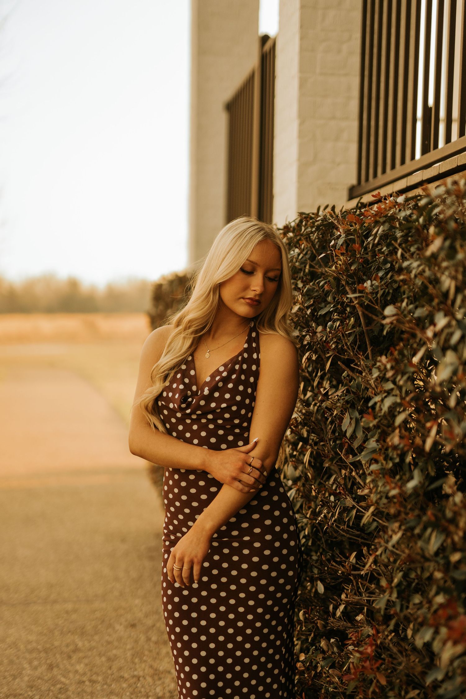 Blonde woman in brown polka dot halter dress leaning against a green hedge wall outdoors.