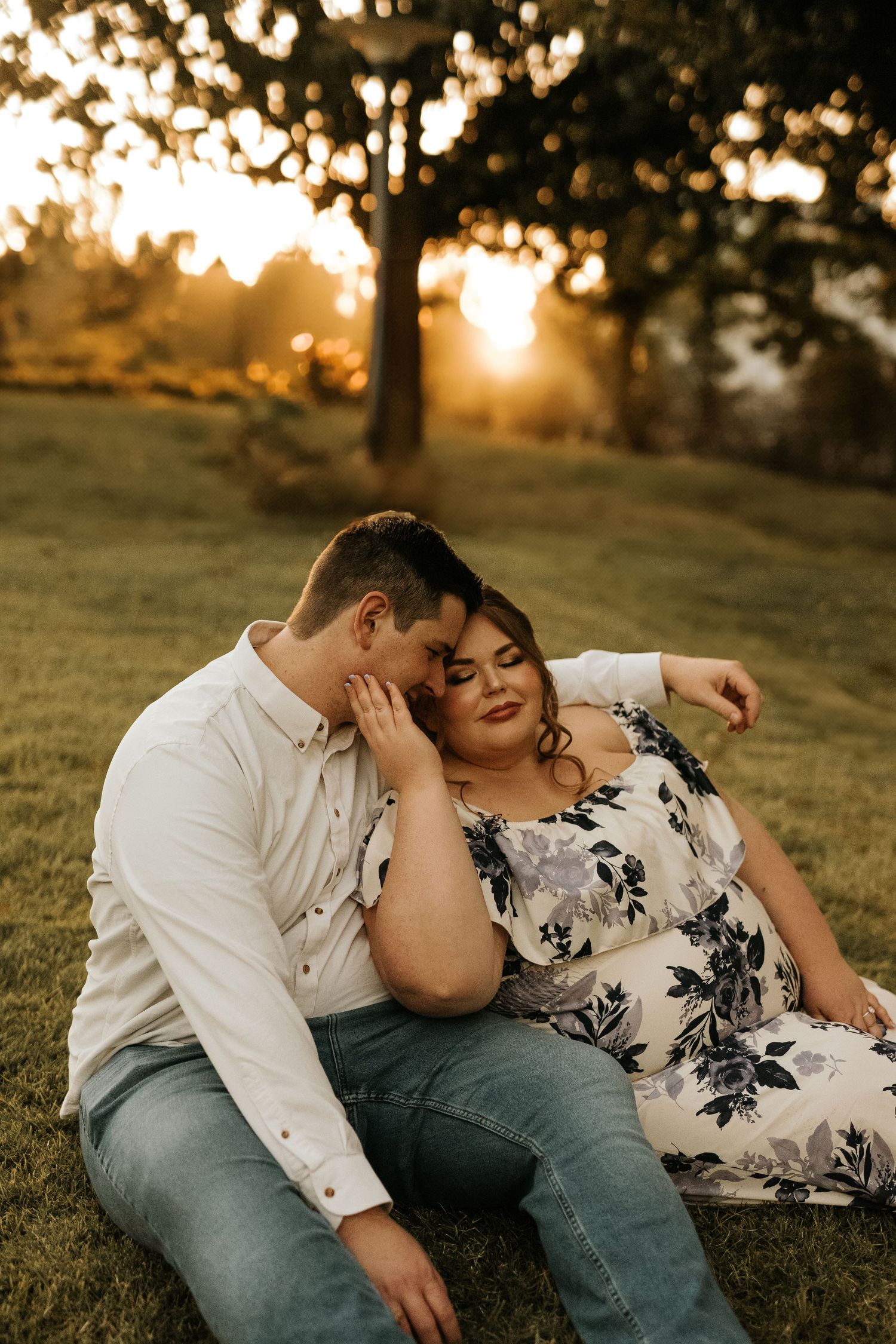Couple sitting on grass at golden hour sunset, woman in floral dress resting on man in white shirt.