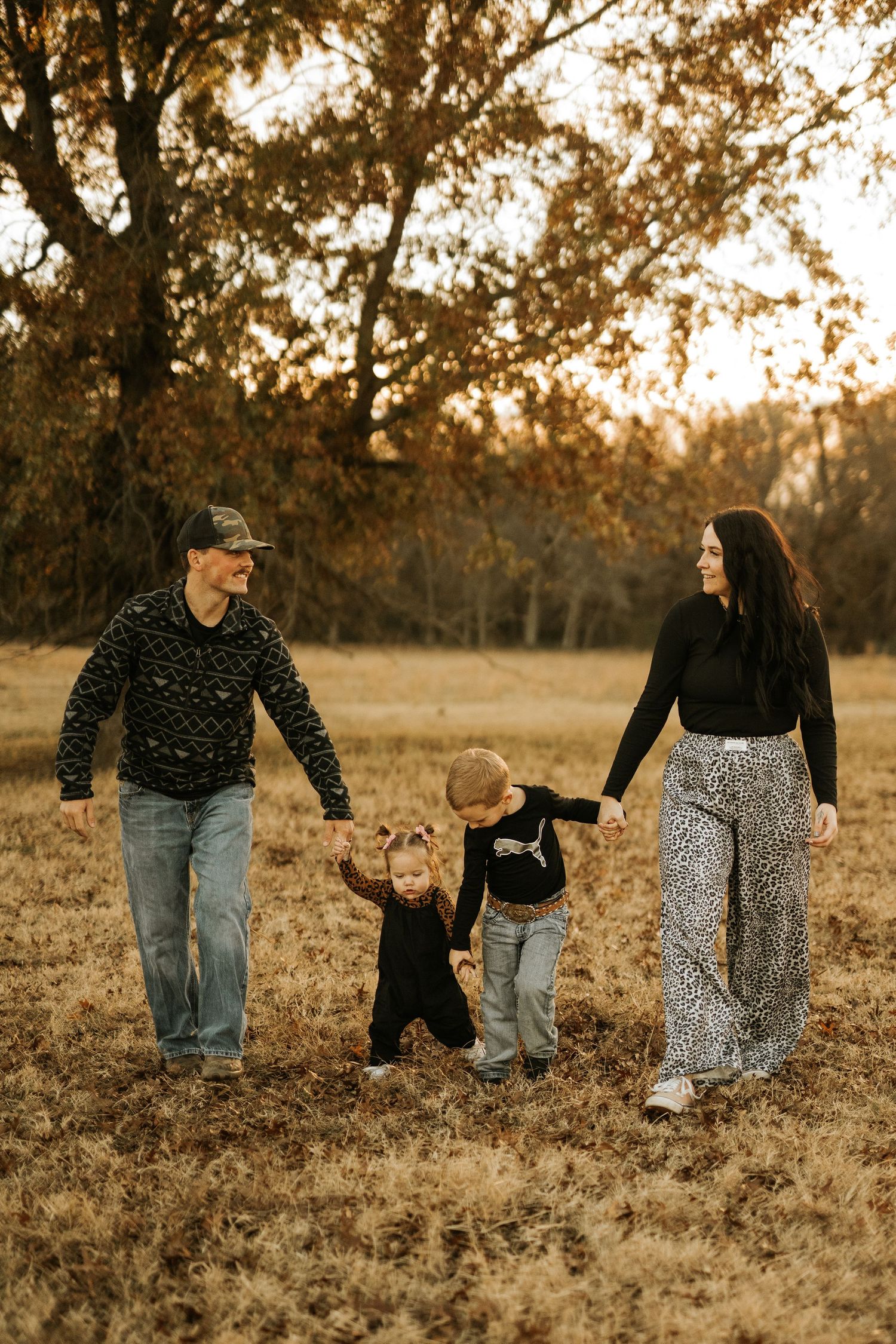 Family of four walking in autumn field at golden hour, parents holding children's hands among fall foliage.