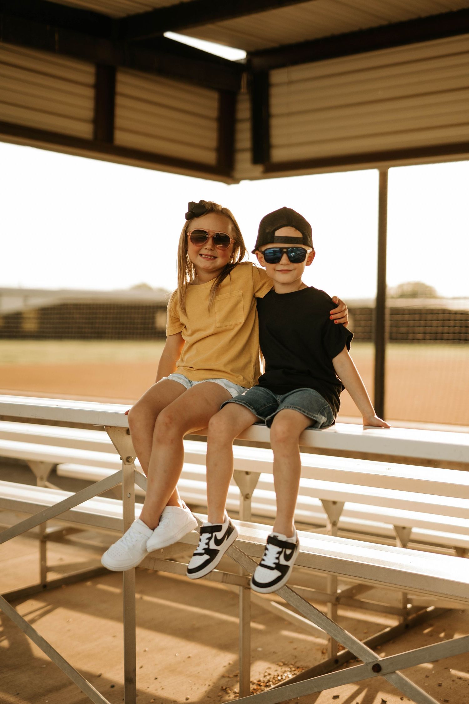 Two kids sitting on bleachers in casual wear and sunglasses.