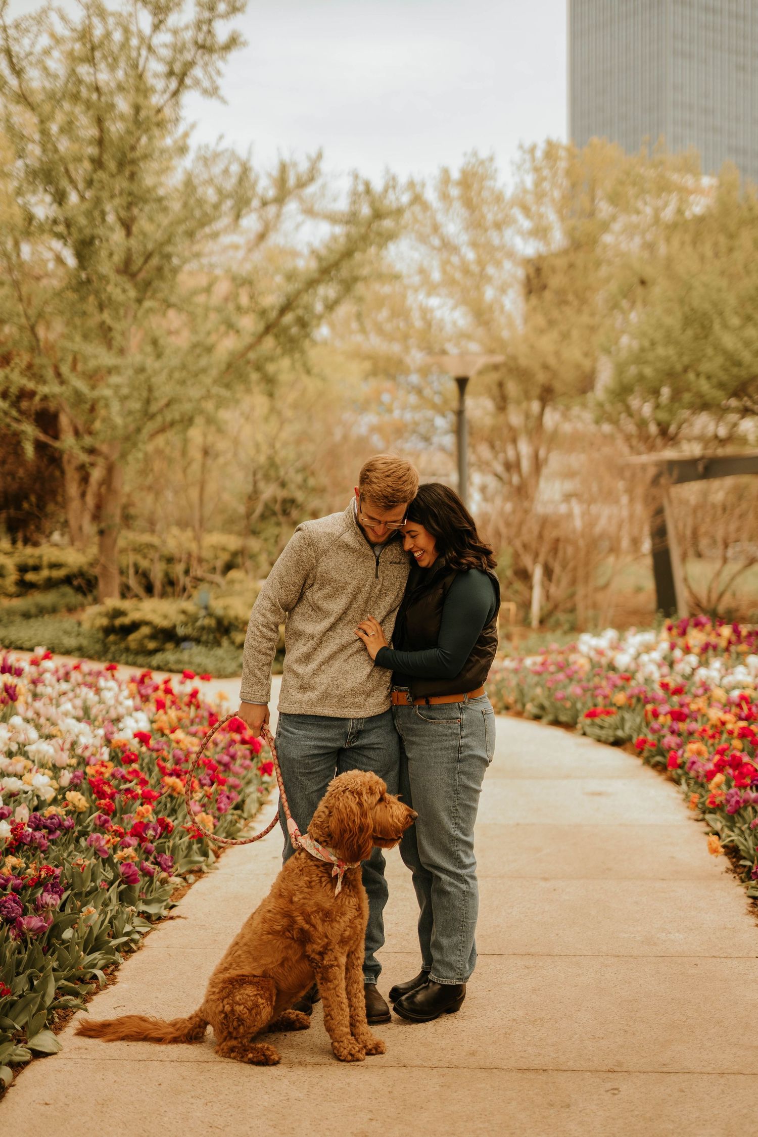 Newly engaged couple celebrates with their golden retriever among blooming tulips.