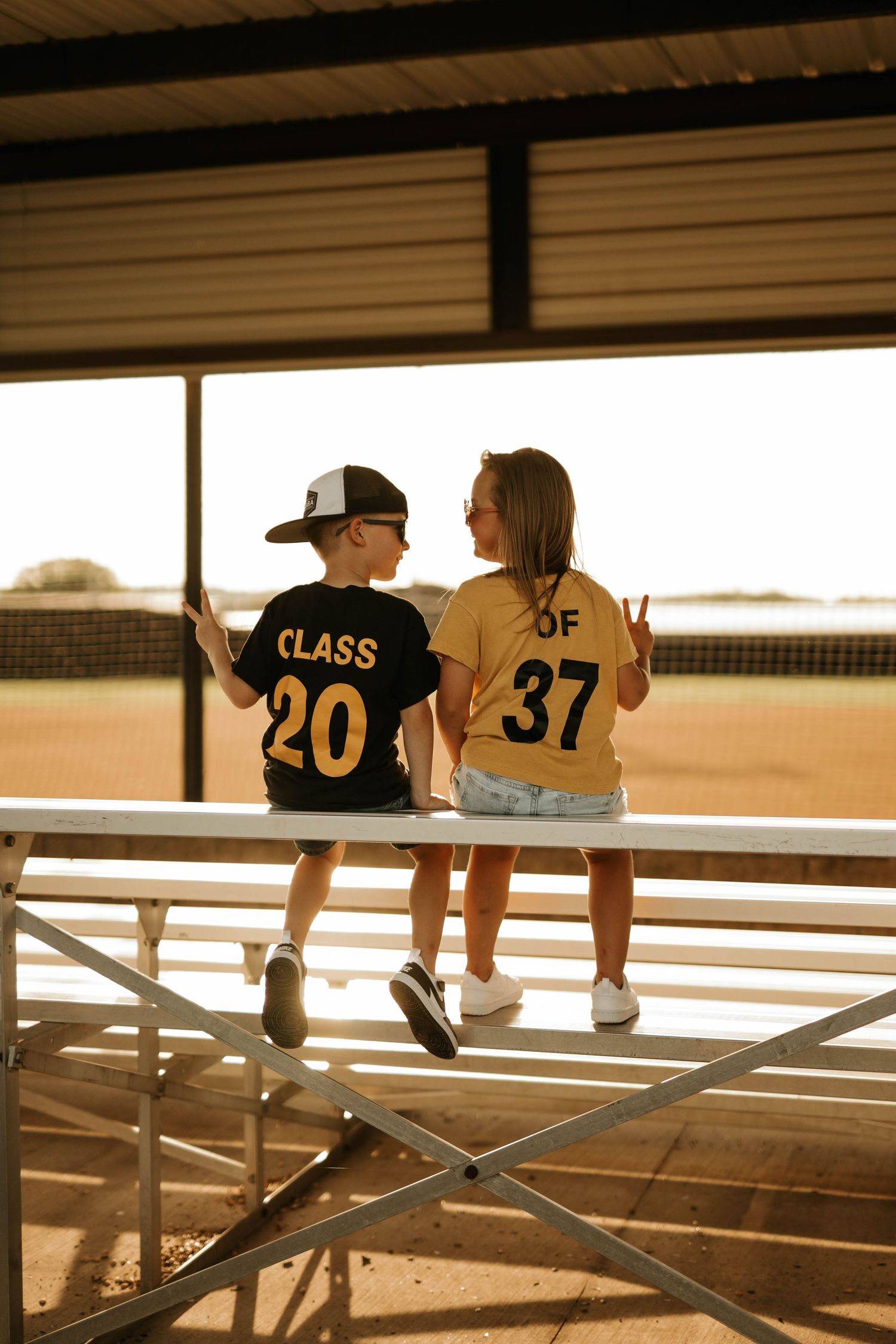 Children wearing numbered jerseys sitting on metal bleachers at sunset.