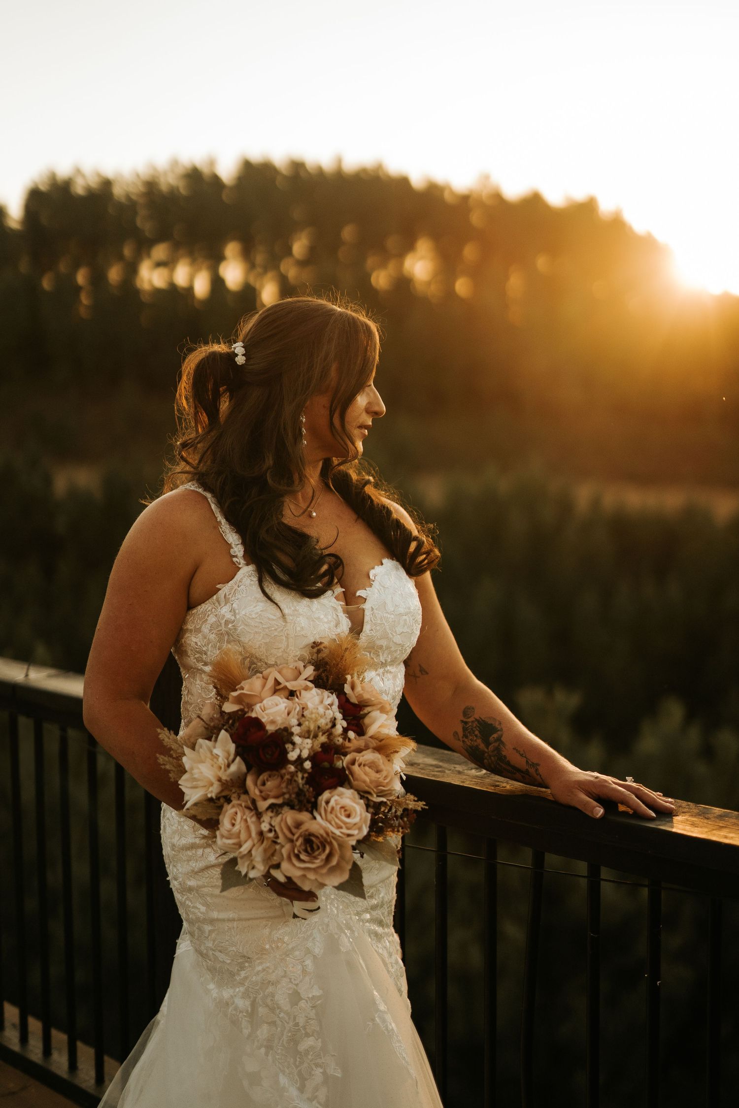 Bride in lace wedding gown holds bouquet of blush roses on a balcony at golden hour sunset.