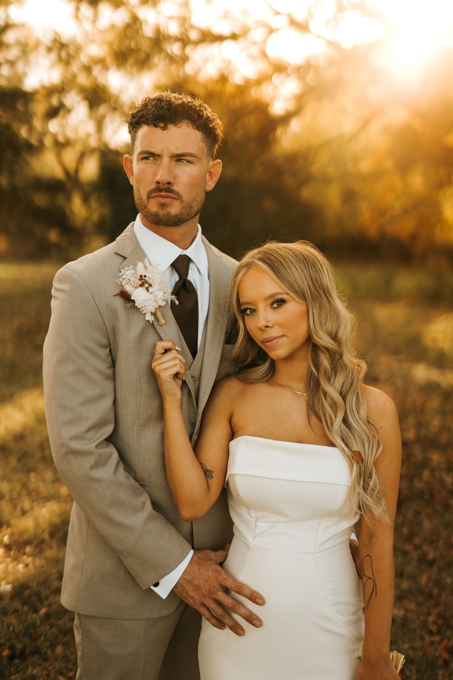 A bride and groom pose together in golden sunset light, she in a white strapless gown, he in a tan suit with boutonniere.