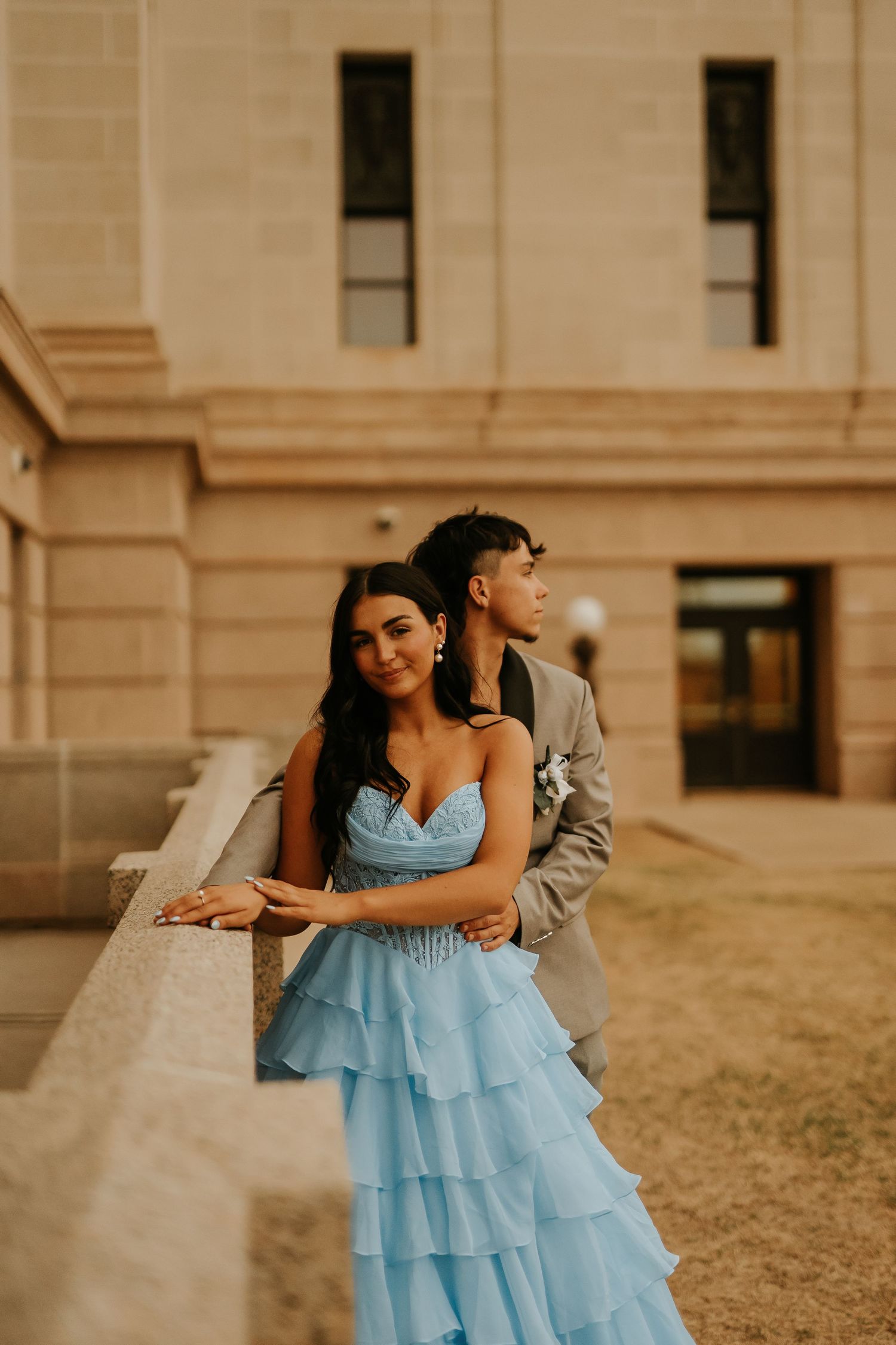 A young couple in prom attire poses outdoors, she in a blue ruffled gown, he in a gray suit, against a stone building.