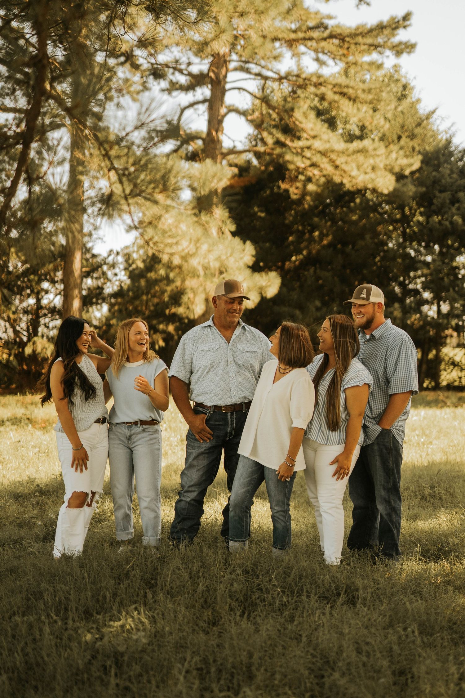A joyful family of six poses together in a sunlit field surrounded by trees, wearing casual white and denim outfits.