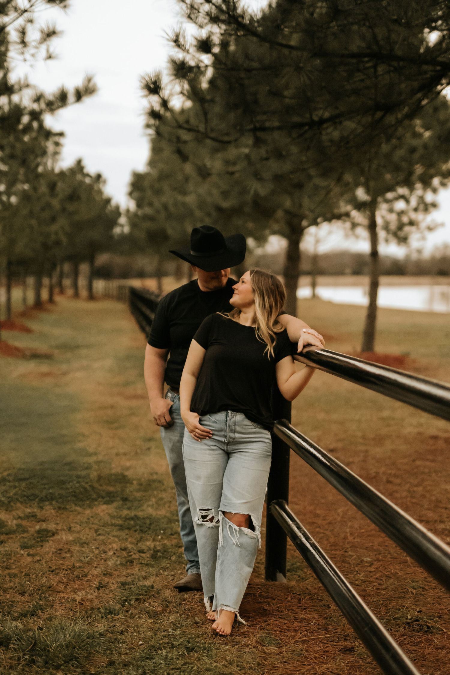 Couple sharing a romantic moment by a rustic fence, man in black cowboy hat and woman in ripped jeans, surrounded by pine trees.