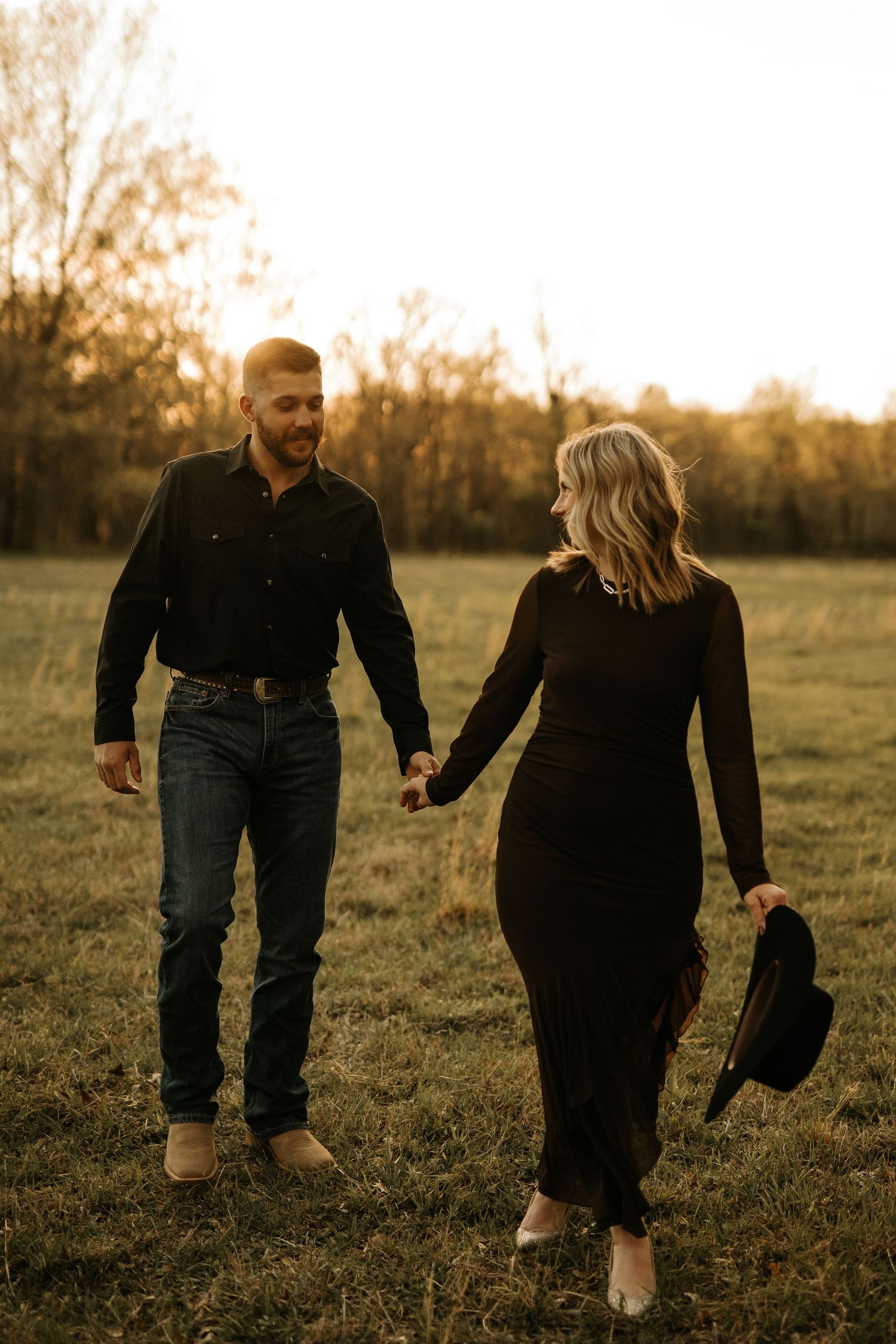 Couple holding hands walking in golden hour field, both dressed in black, woman carrying hat, trees in background.