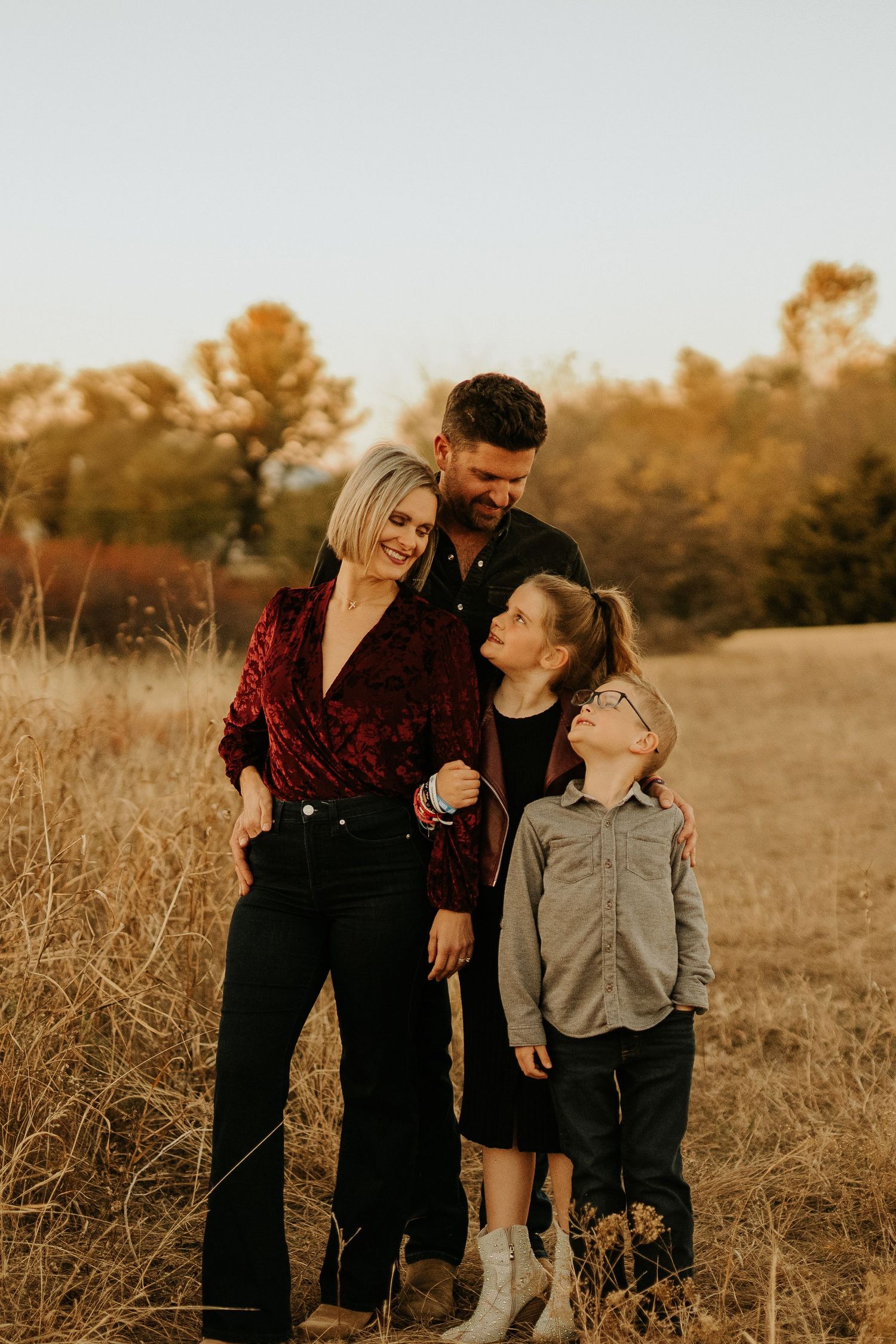 Family of four posing in golden autumn field, parents embracing two children, warm fall foliage backdrop.