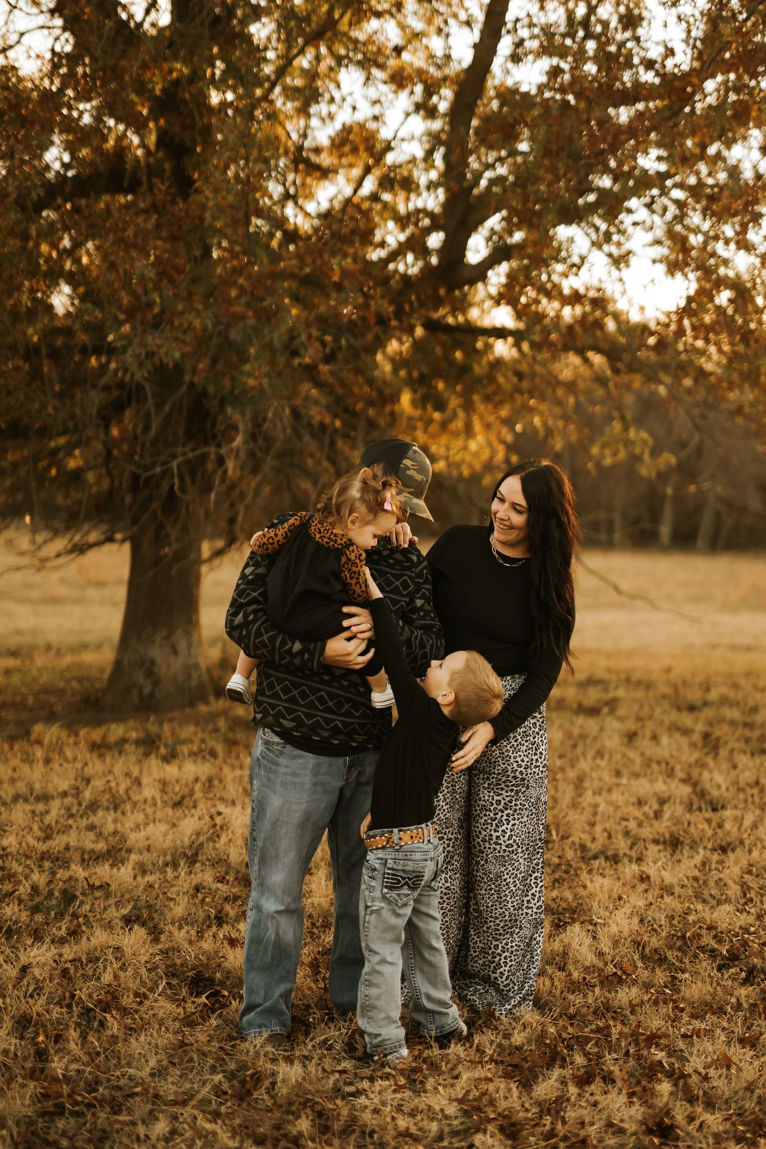 Family of four enjoying a golden autumn day outdoors, with warm fall foliage and a large oak tree in the background.