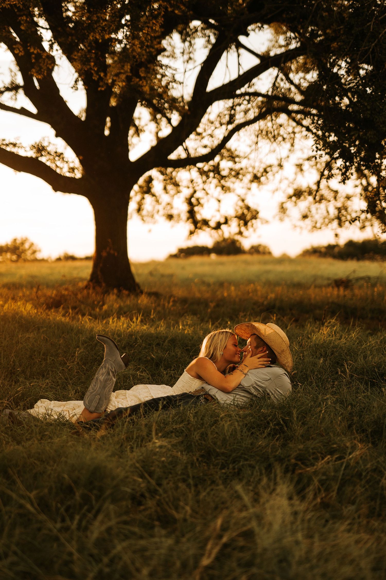 Couple lying in golden grass beneath a large oak tree at sunset, sharing a romantic embrace outdoors.