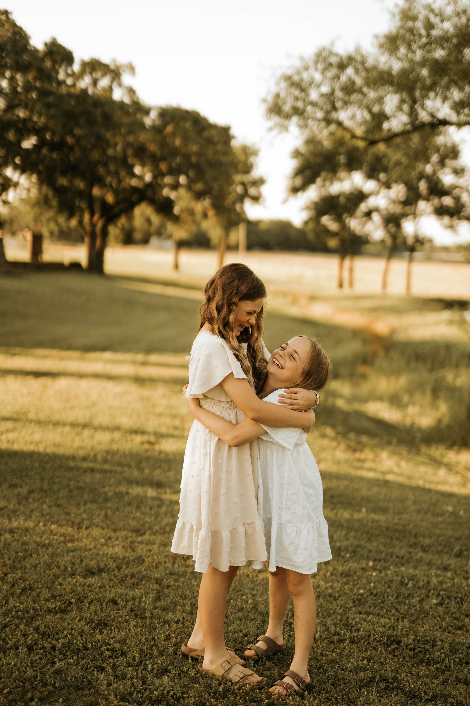 Two children in white dresses share a joyful embrace on a grassy lawn.