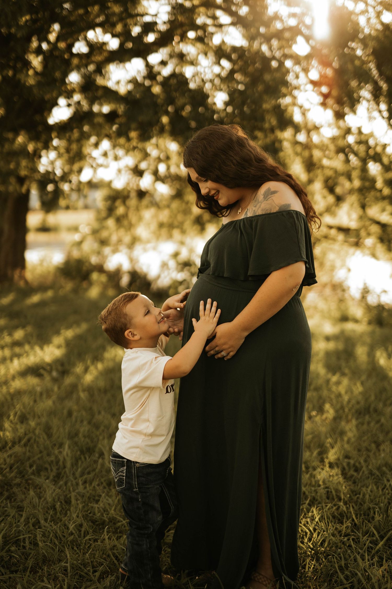 A pregnant woman in a black dress shares a tender moment with her young son in a sunlit outdoor setting.
