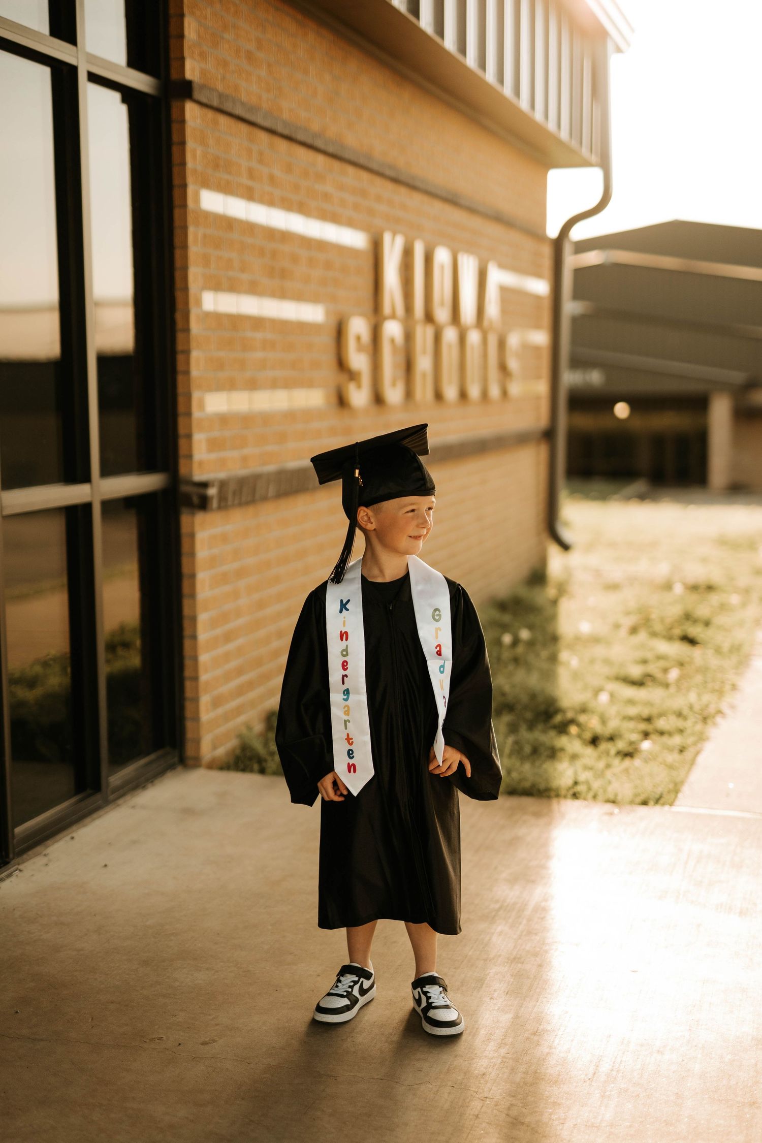 Graduate in black gown and cap stands in front of brick school building wearing custom sash.