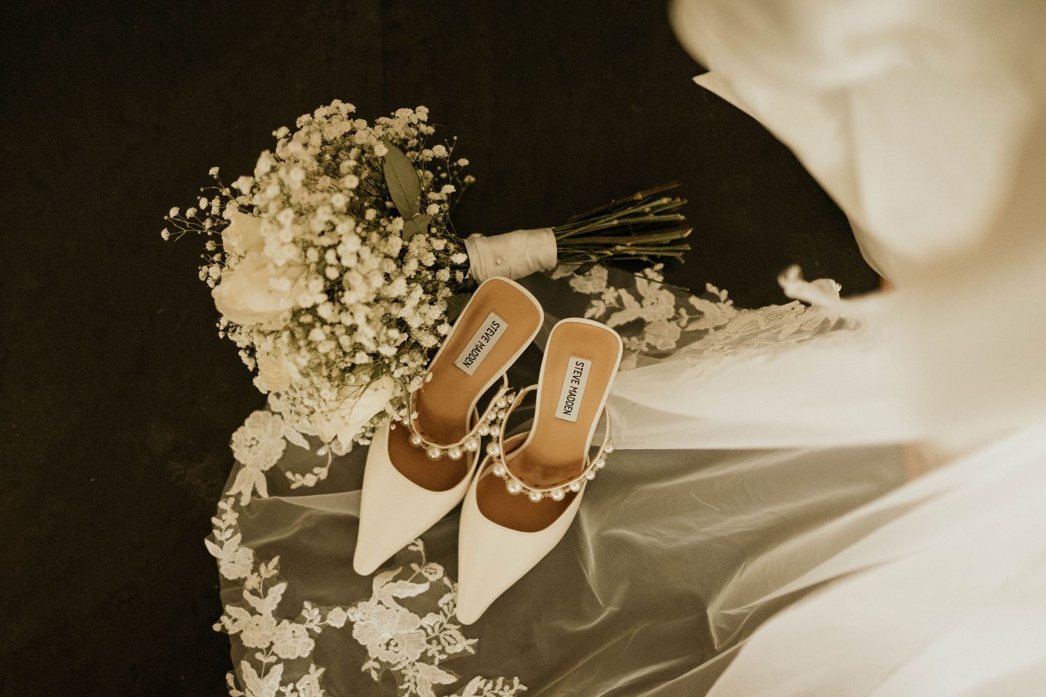 Elegant white bridal shoes and baby's breath bouquet arranged on dark background.