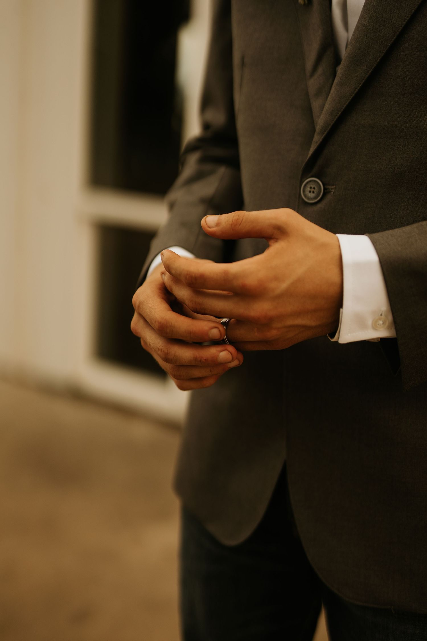 A man in a dark gray suit adjusts his cufflinks, wearing a wedding ring on his finger.