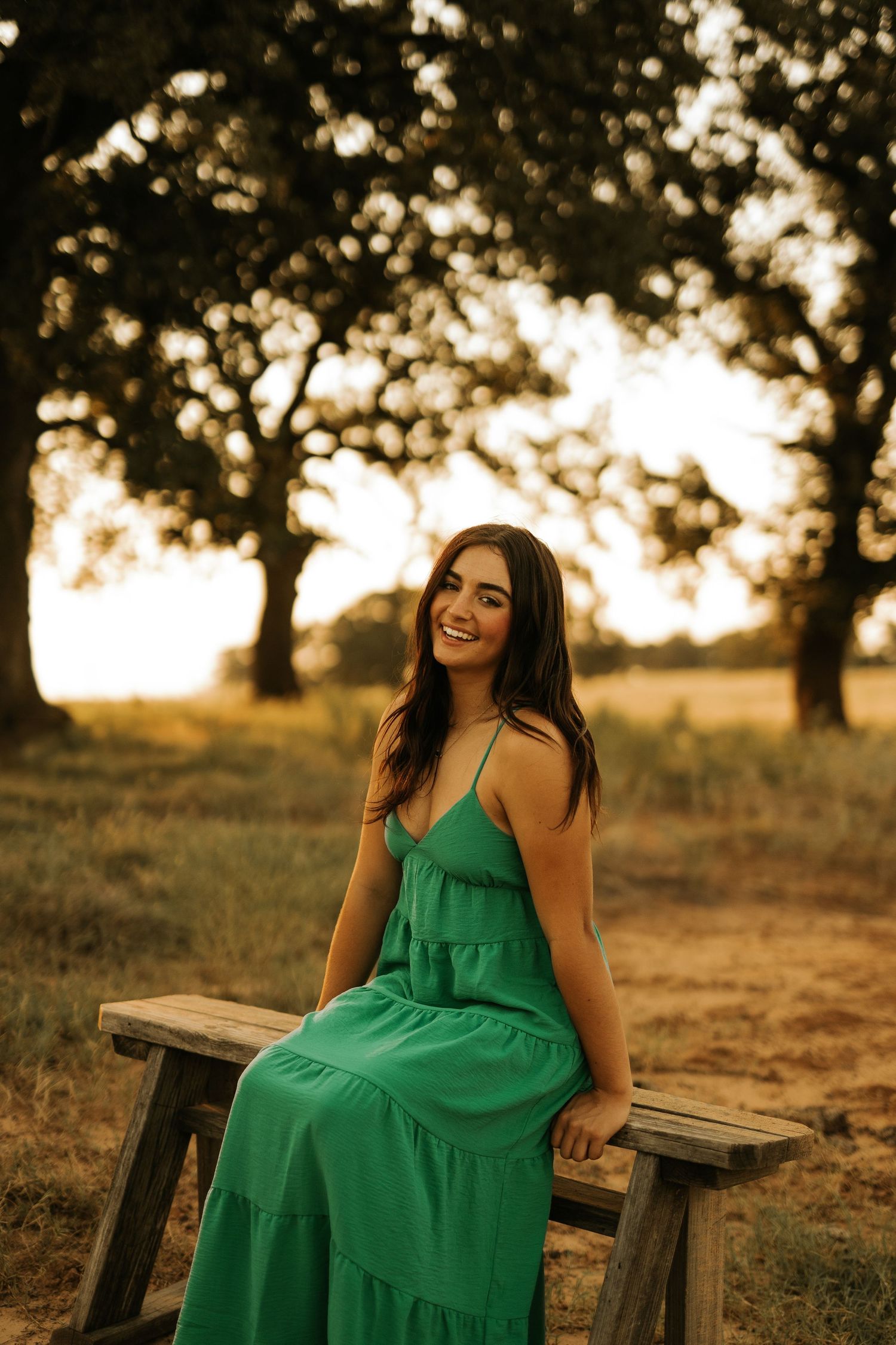 A young woman in a green maxi dress sits on a wooden bench, smiling outdoors at golden hour.
