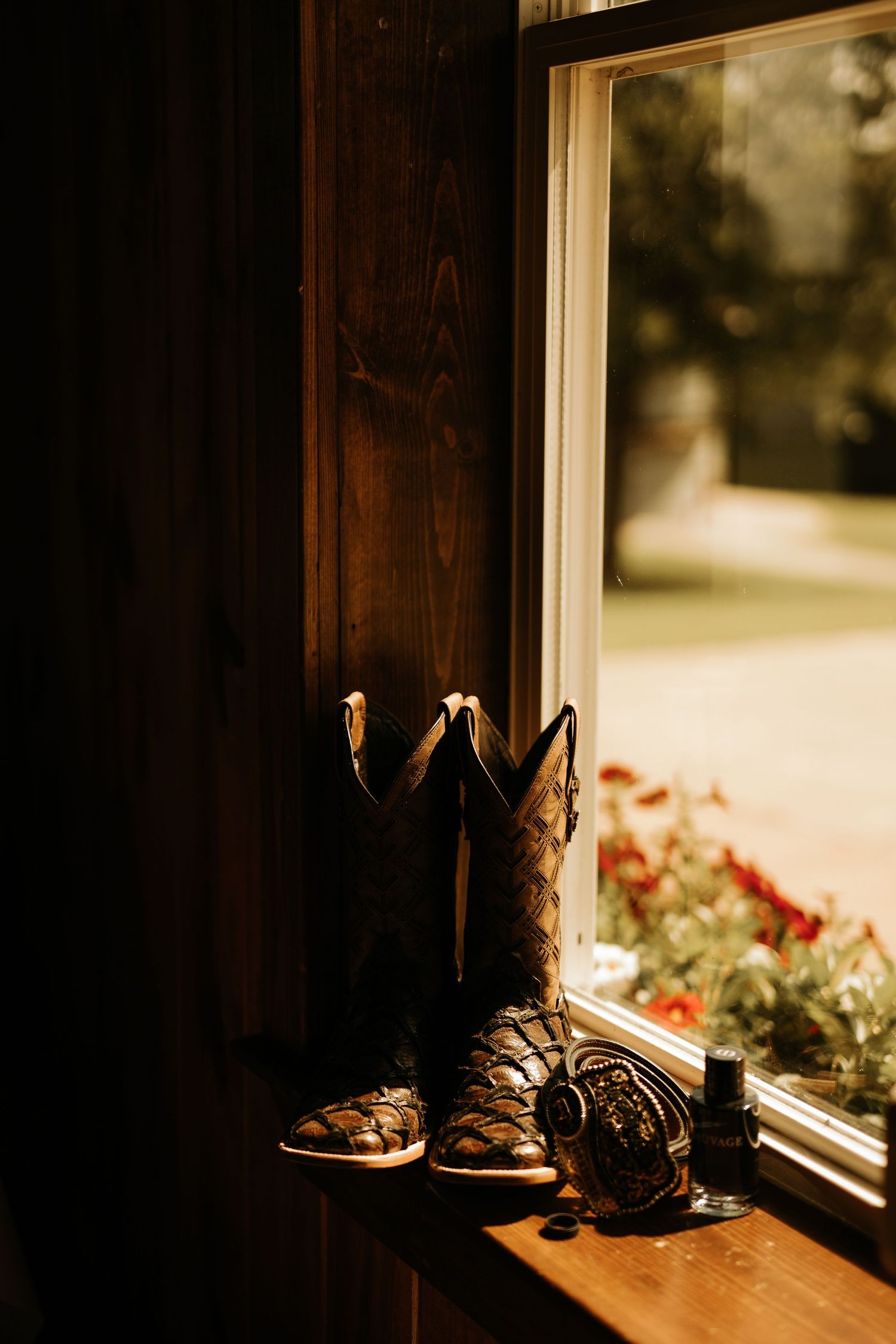 Cowboy boots and accessories on a rustic wooden windowsill with warm golden light and a garden view outside.