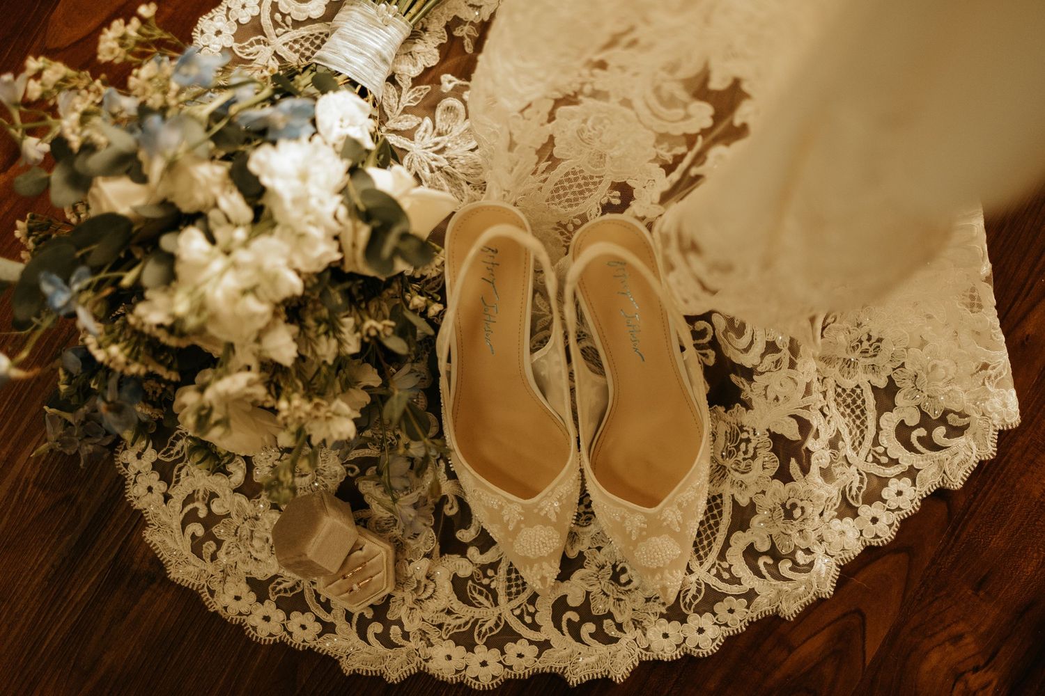 Elegant bridal heels with lace detailing rest on a wedding dress beside a blue and white floral bouquet.