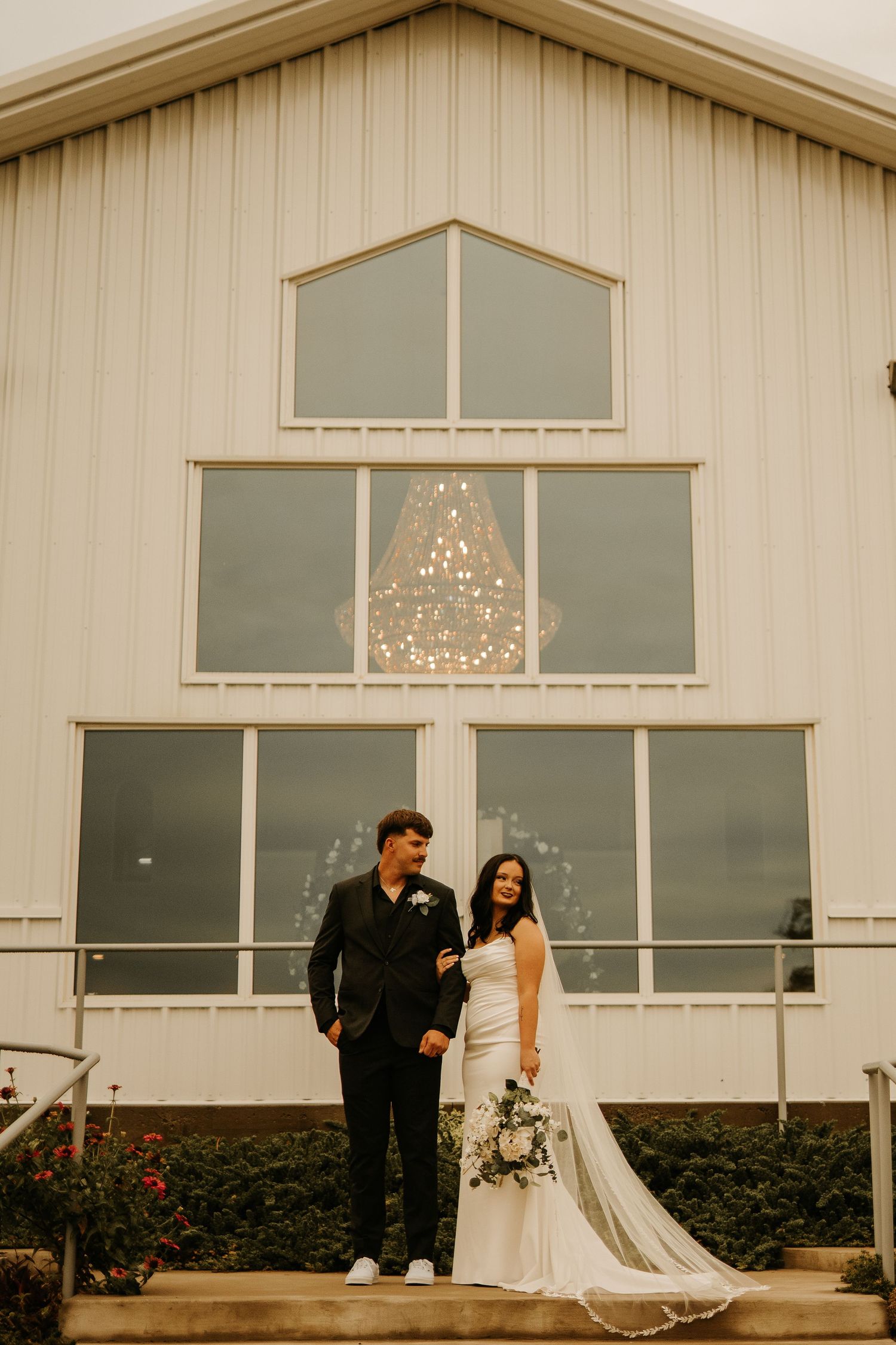 A bride and groom pose together in front of a white barn wedding venue with large windows and a chandelier.