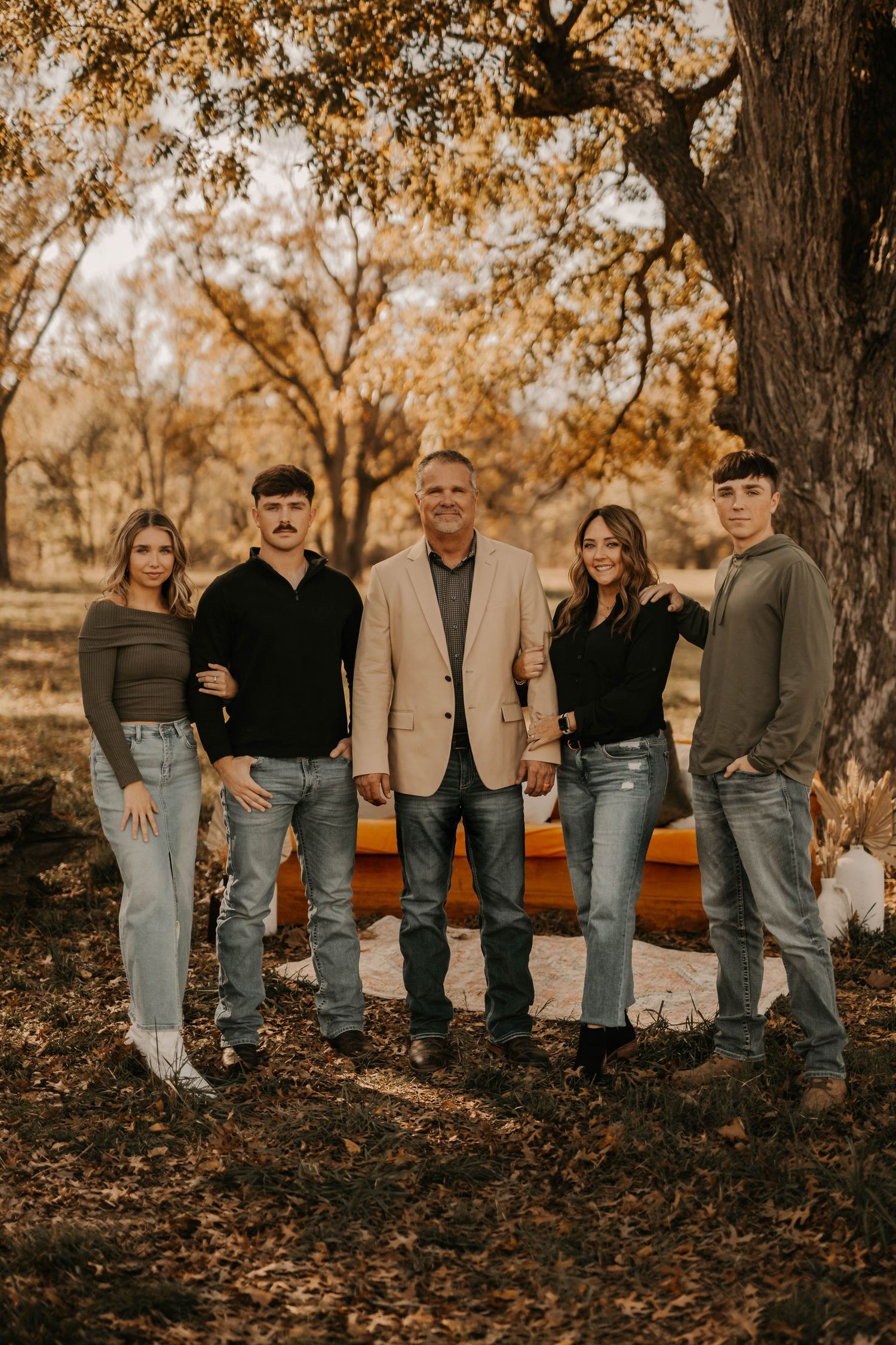Family of five posing outdoors among autumn trees, wearing casual jeans and jackets for a fall family portrait.