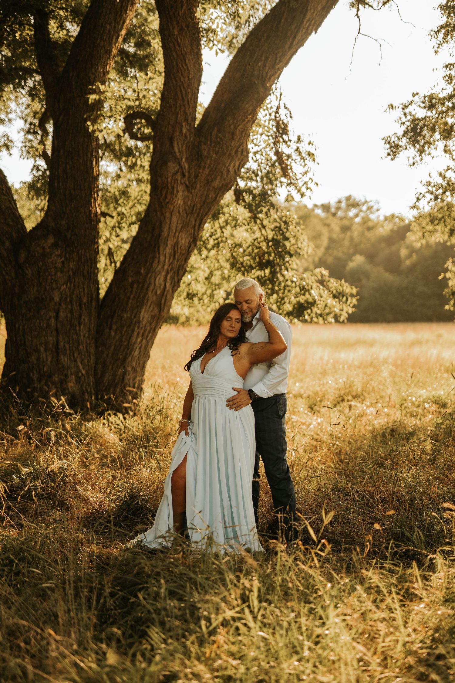 Couple embracing under a large tree in a golden field, woman in flowing white dress, maternity photo session at sunset.