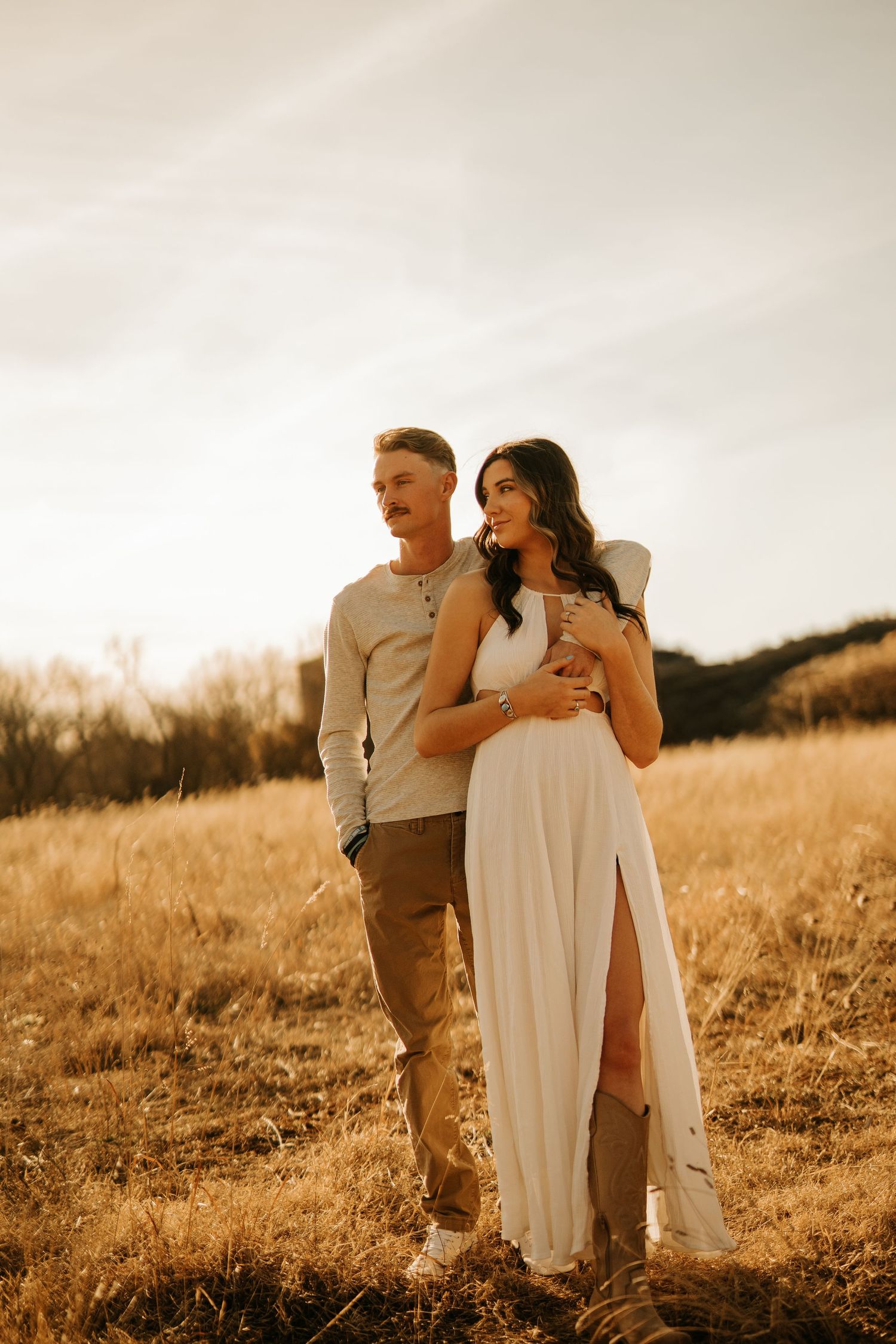 Couple embracing in golden field at sunset, woman in white dress, man in beige shirt and tan pants.