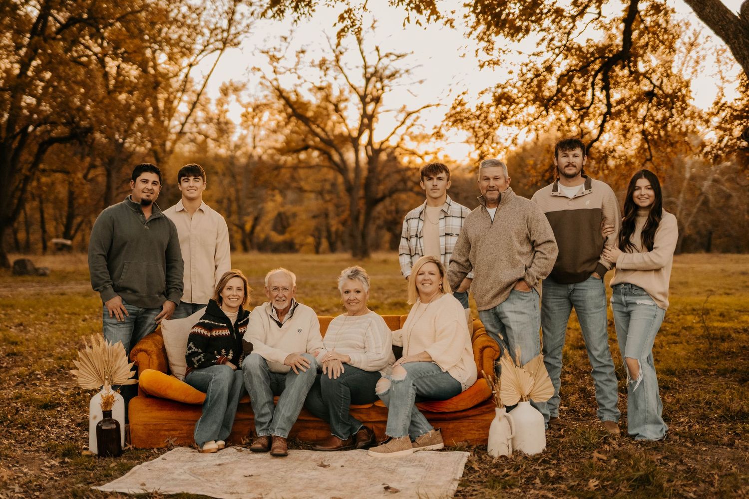 Family of nine poses outdoors among autumn trees during golden hour with pumpkins and a blanket.