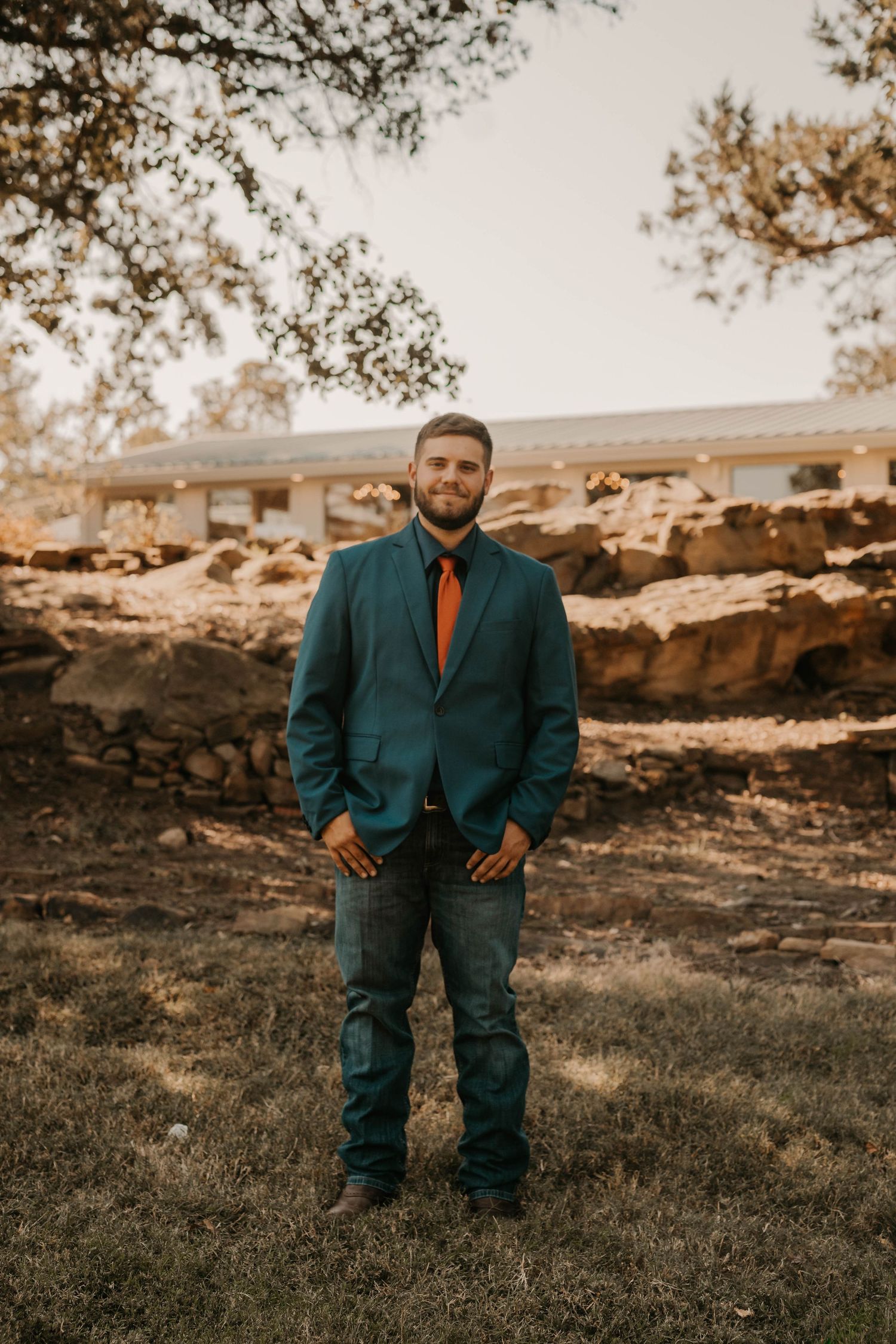 A groom in a teal suit jacket and orange tie poses outdoors on autumn day.