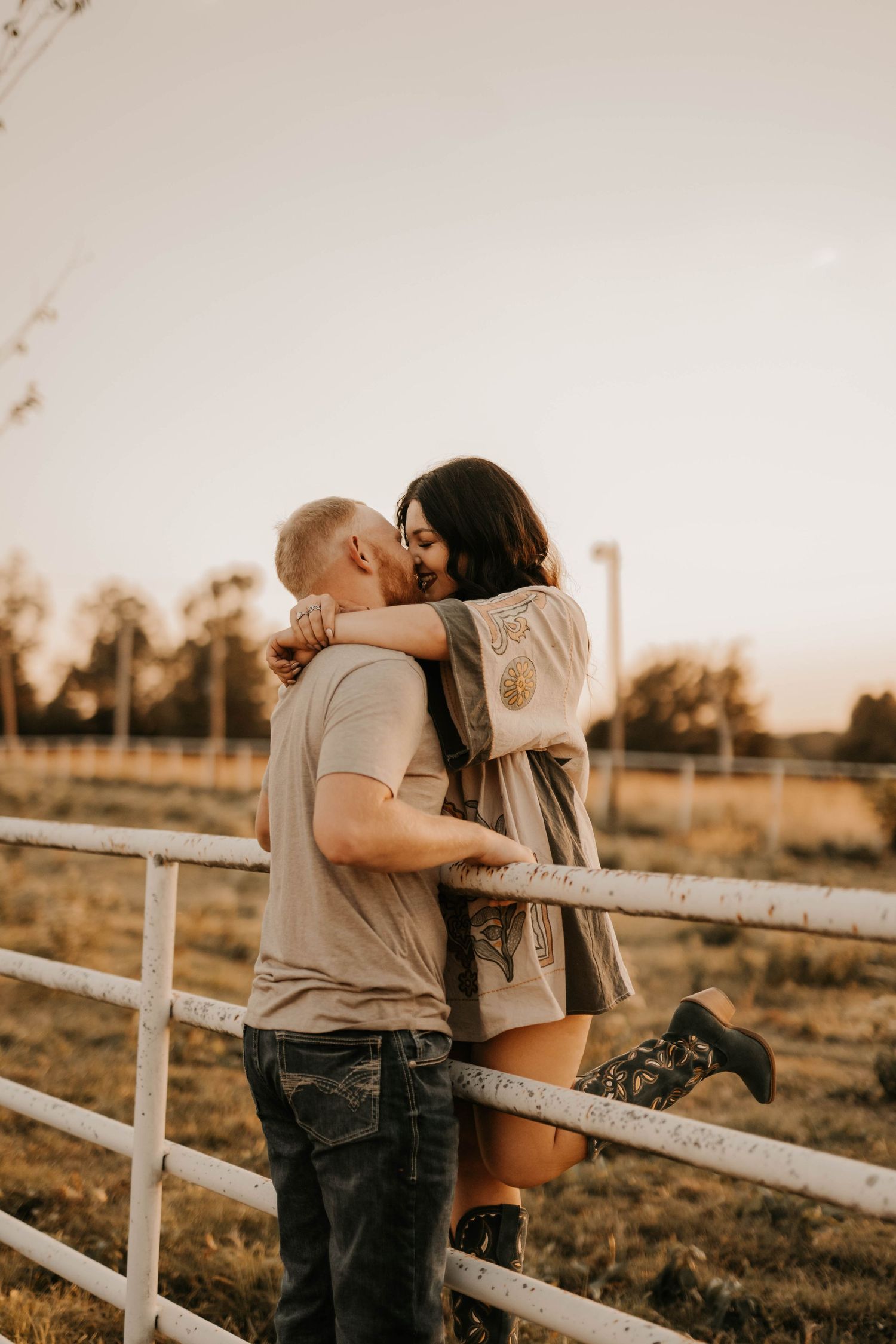 Romantic couple embracing by a white fence during sunset in a rural setting.