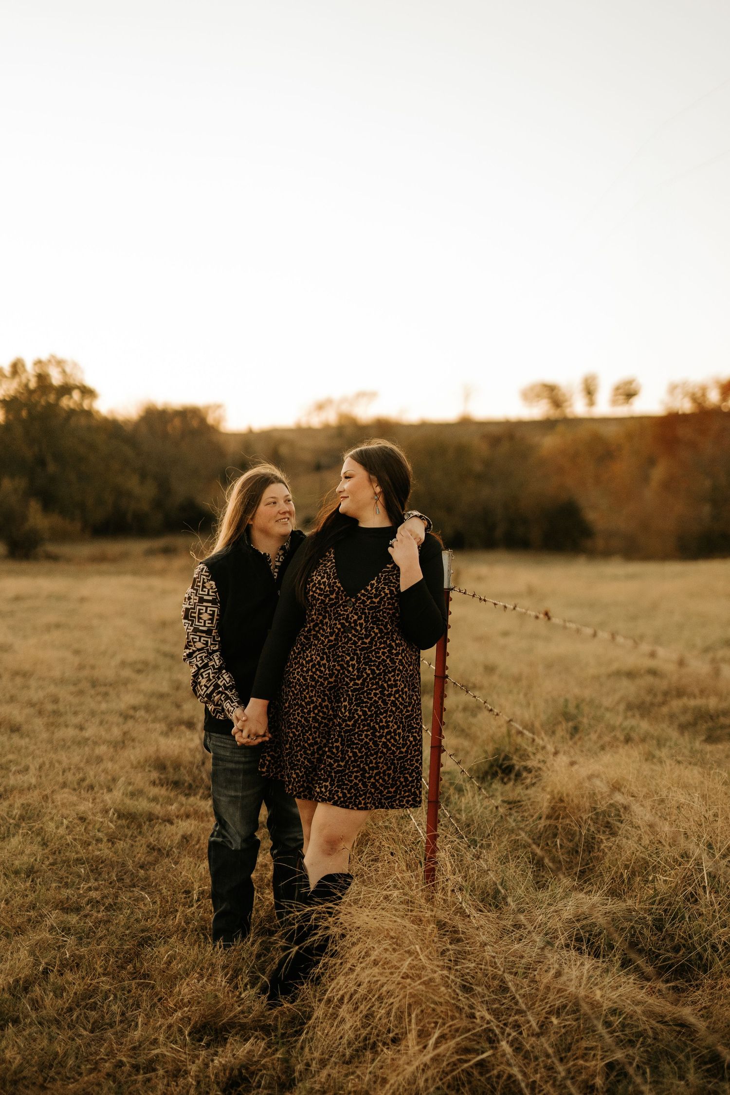 Two women hold hands in a golden field at sunset, smiling at each other near a rustic fence.