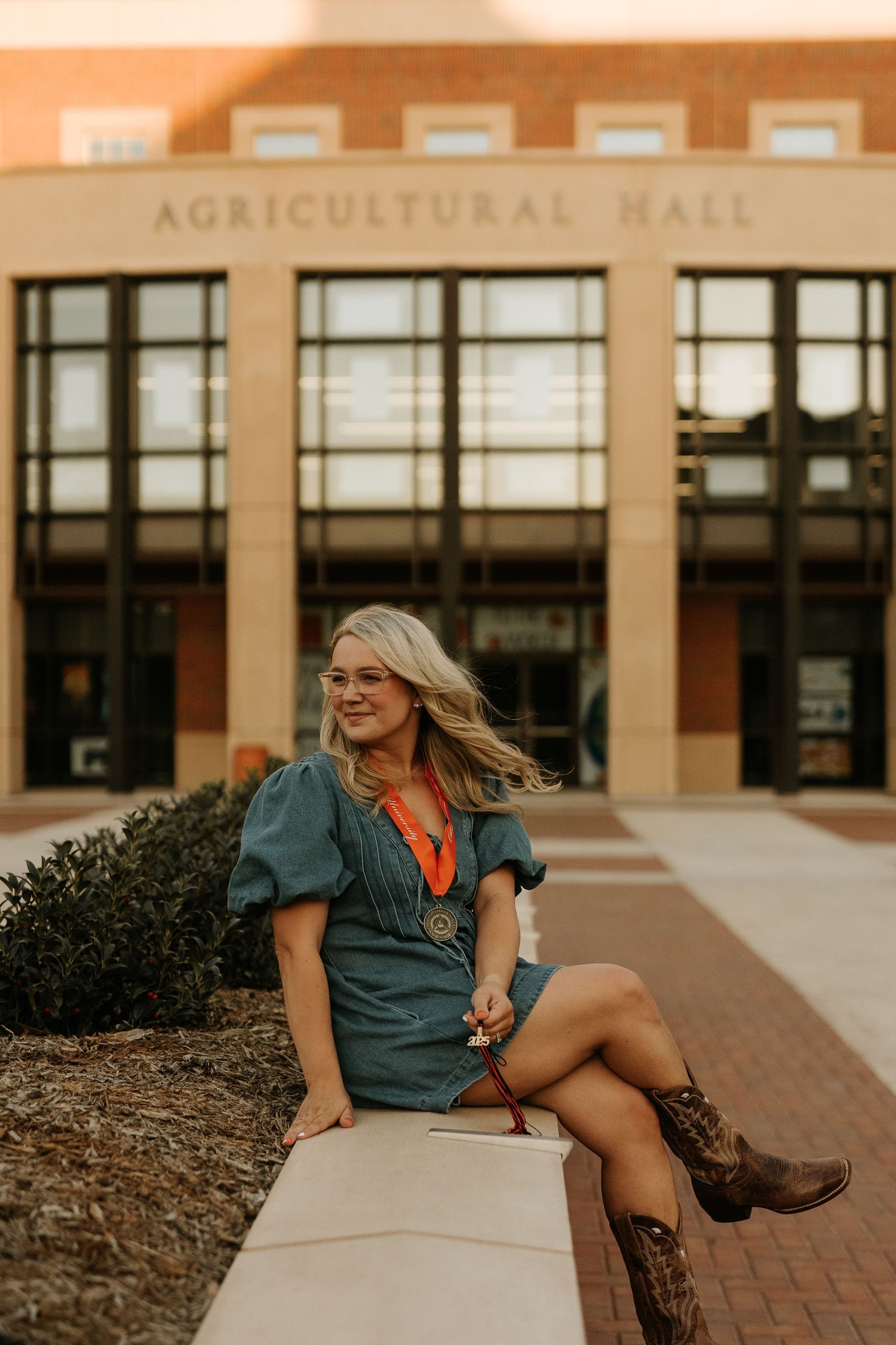 Young woman in teal dress and cowboy boots sitting outside Agricultural Hall building on a sunny day.
