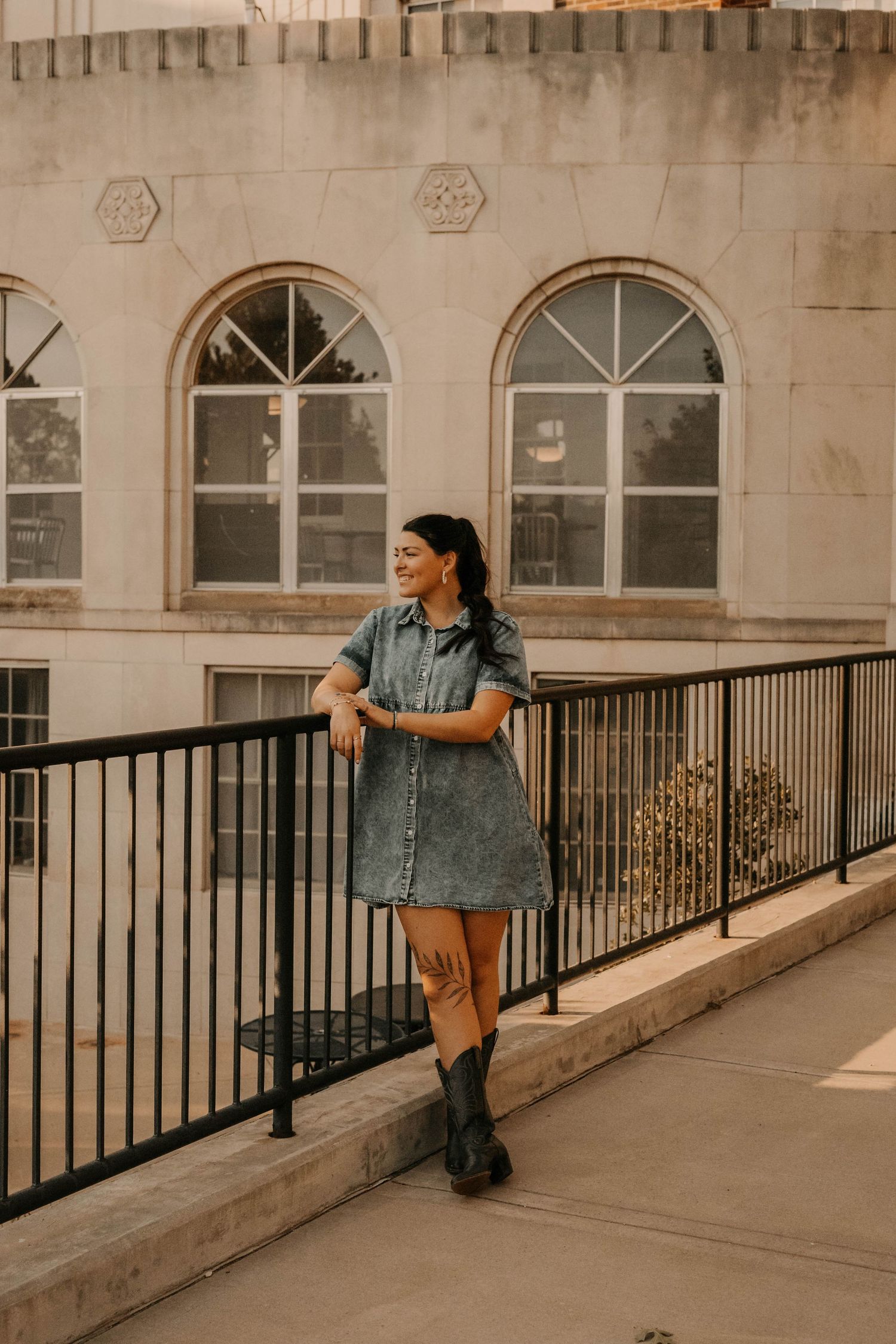 Person in denim dress stands against railing of historic building with arched windows.