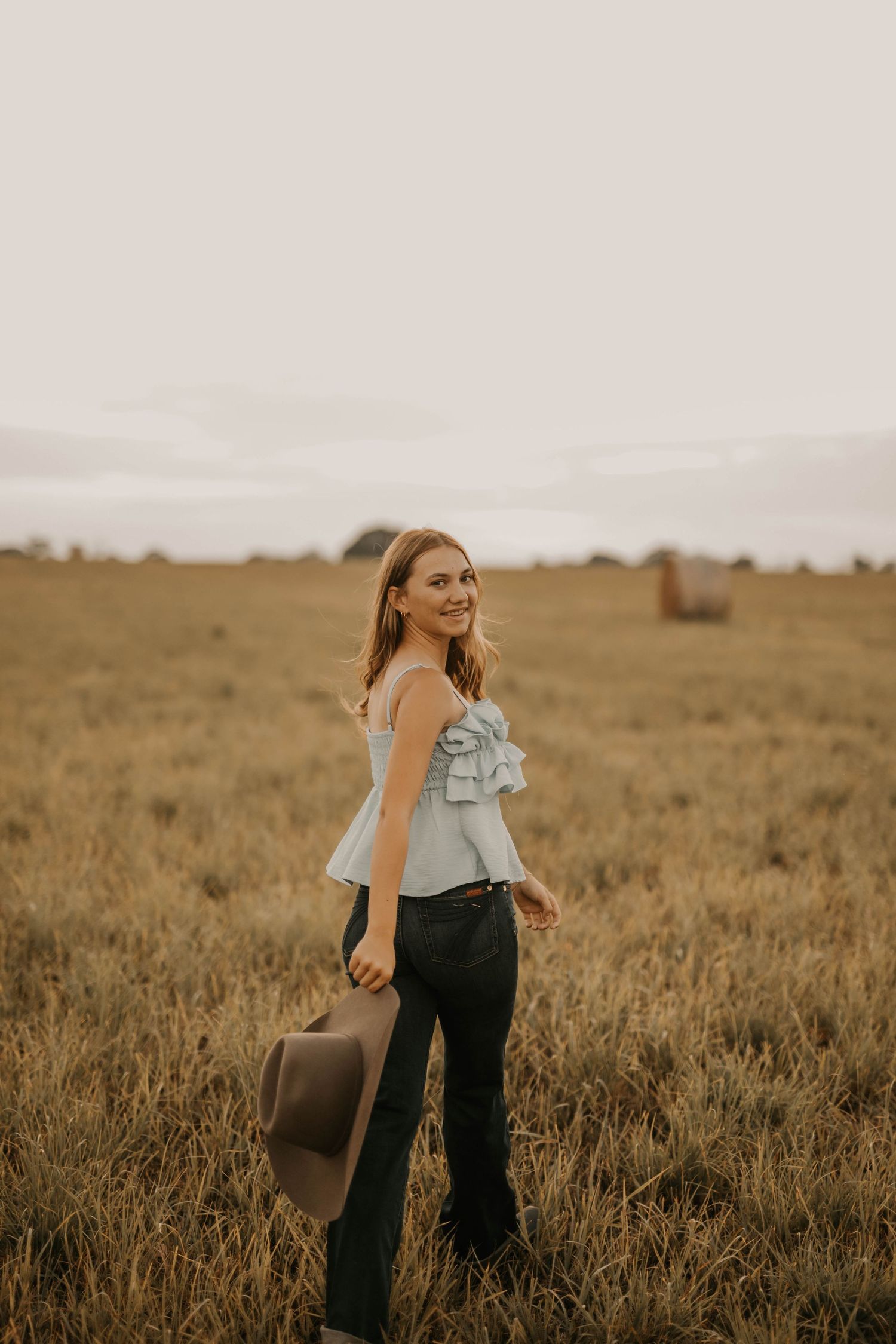 Woman in ruffled top poses in open field at sunset holding wide-brim hat.