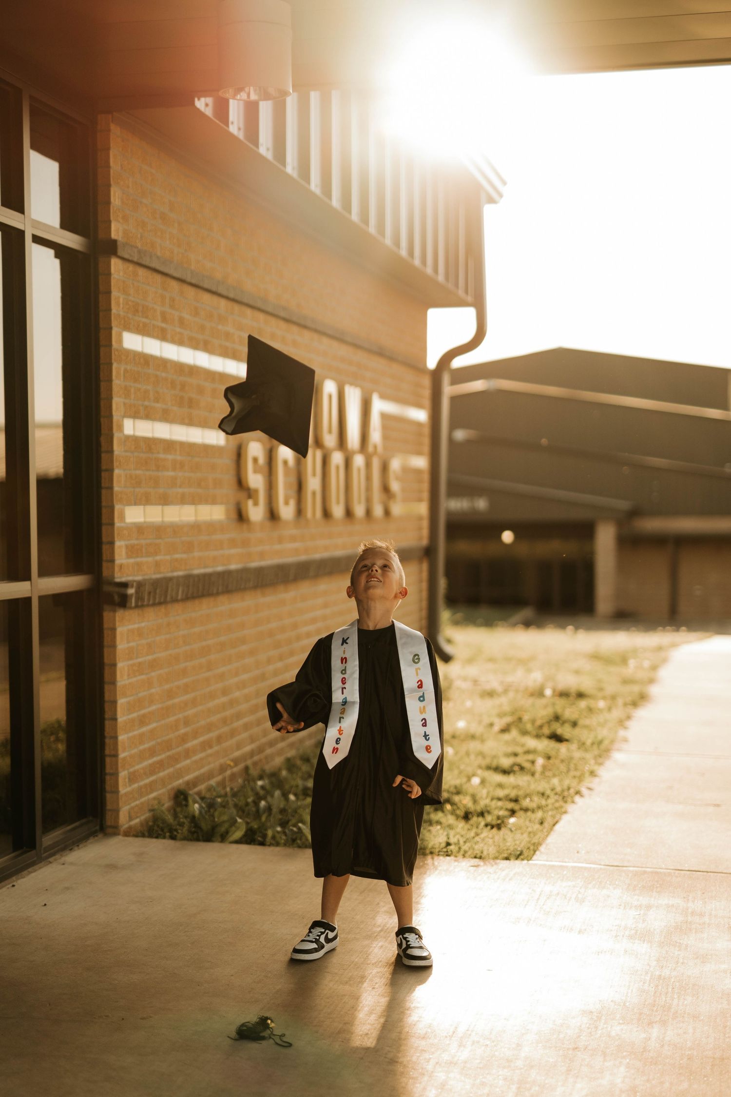 Sunlit graduation portrait of student in black gown and mortarboard outside school entrance.