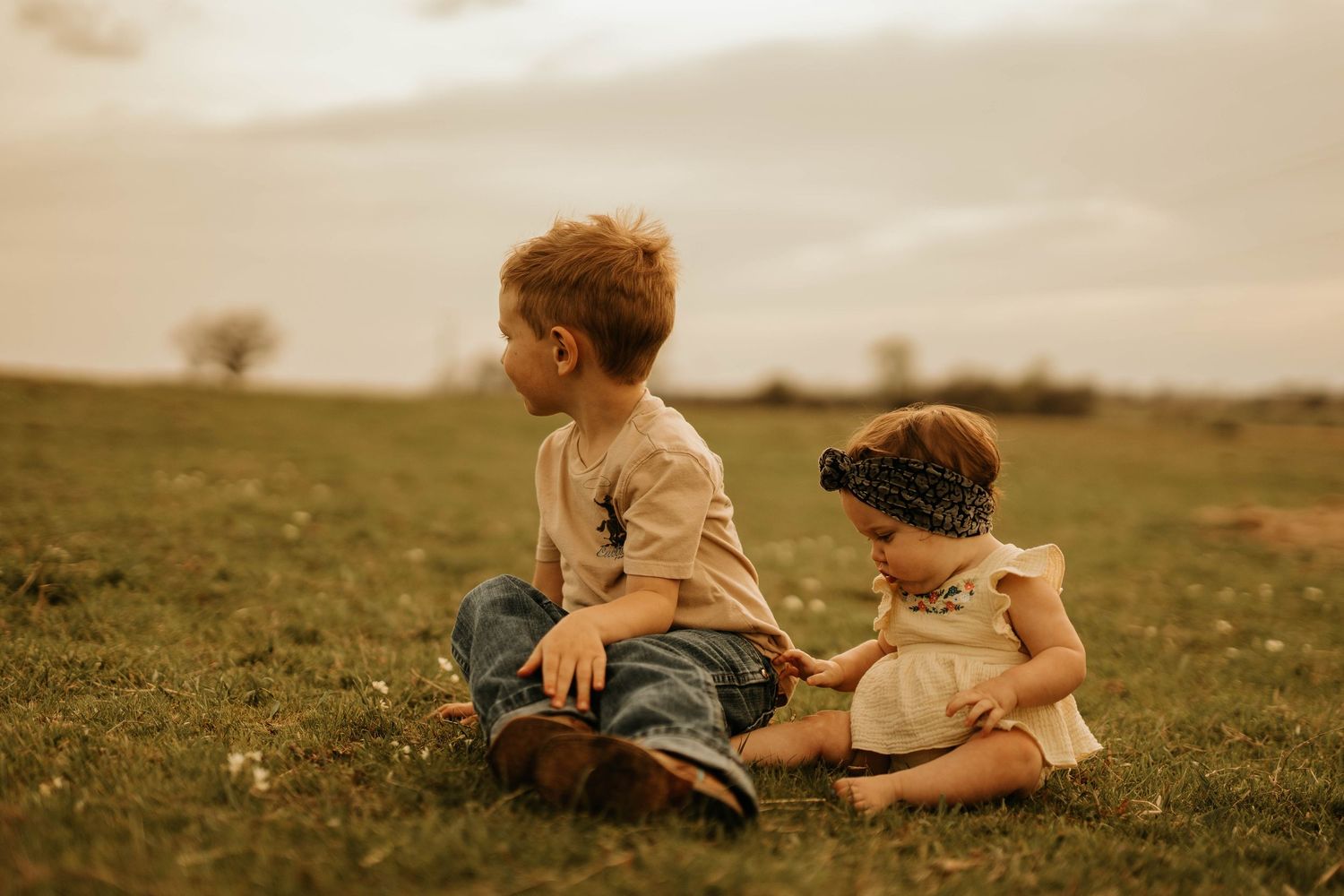 Two young children sit together on grassy field during sunset in vintage-toned photograph.