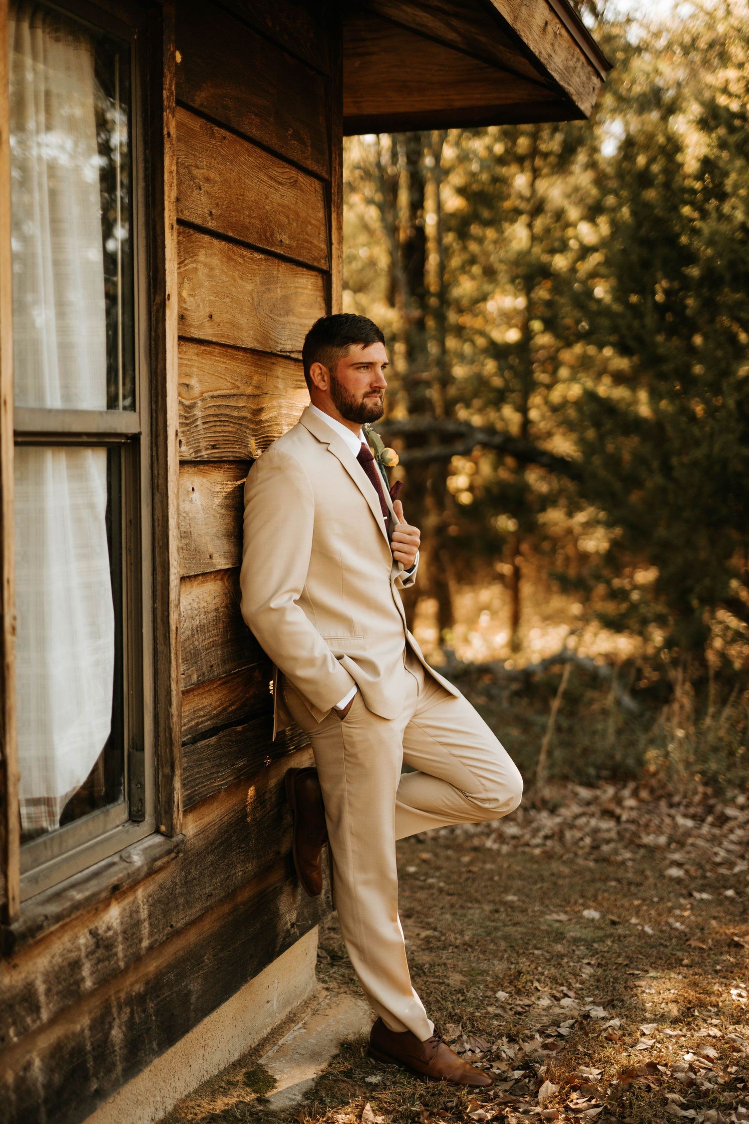 A well-dressed figure leans against a rustic wooden cabin in warm evening light.