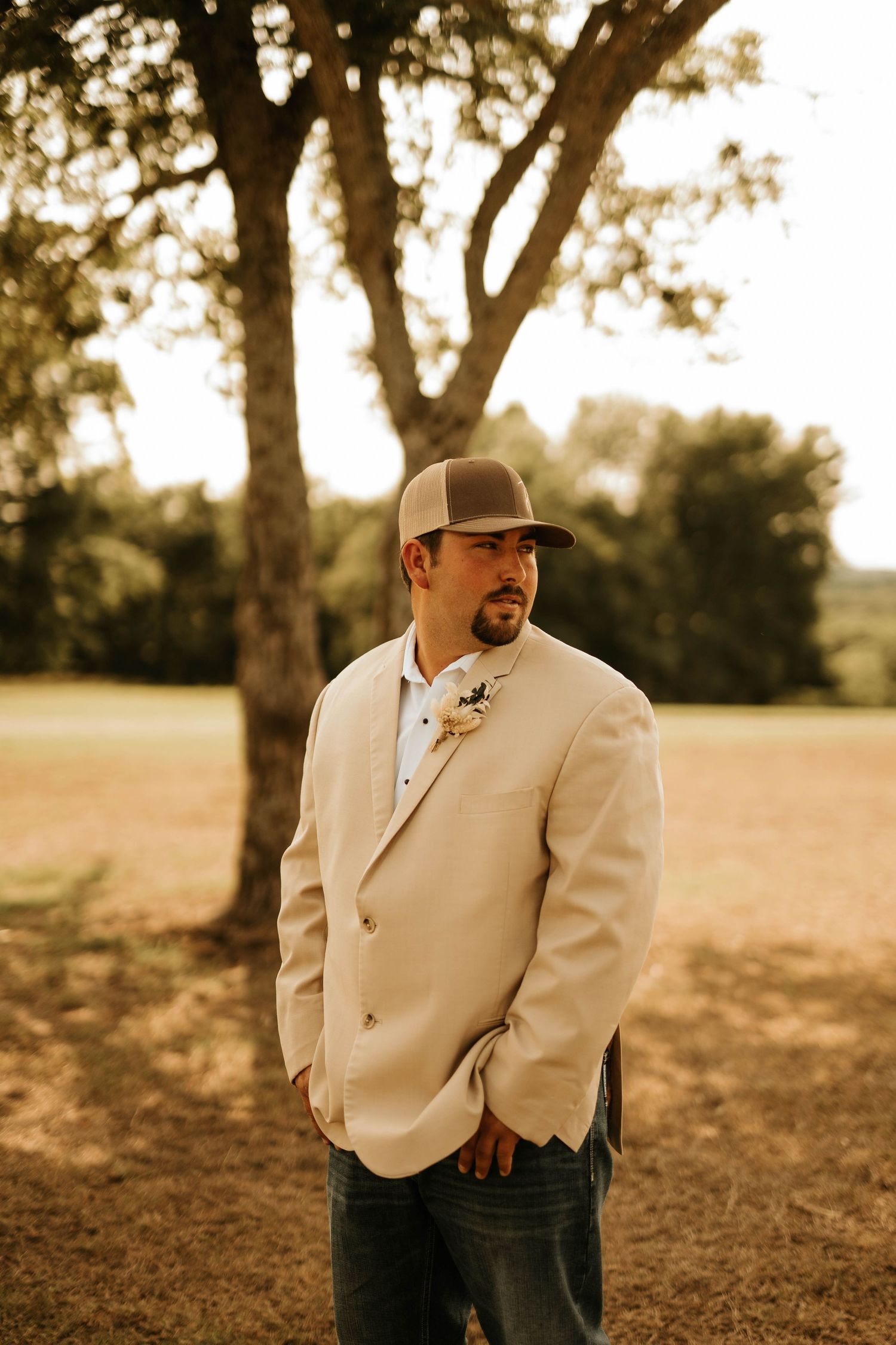 A groom in a beige suit and cowboy hat stands in a sun-drenched field.
