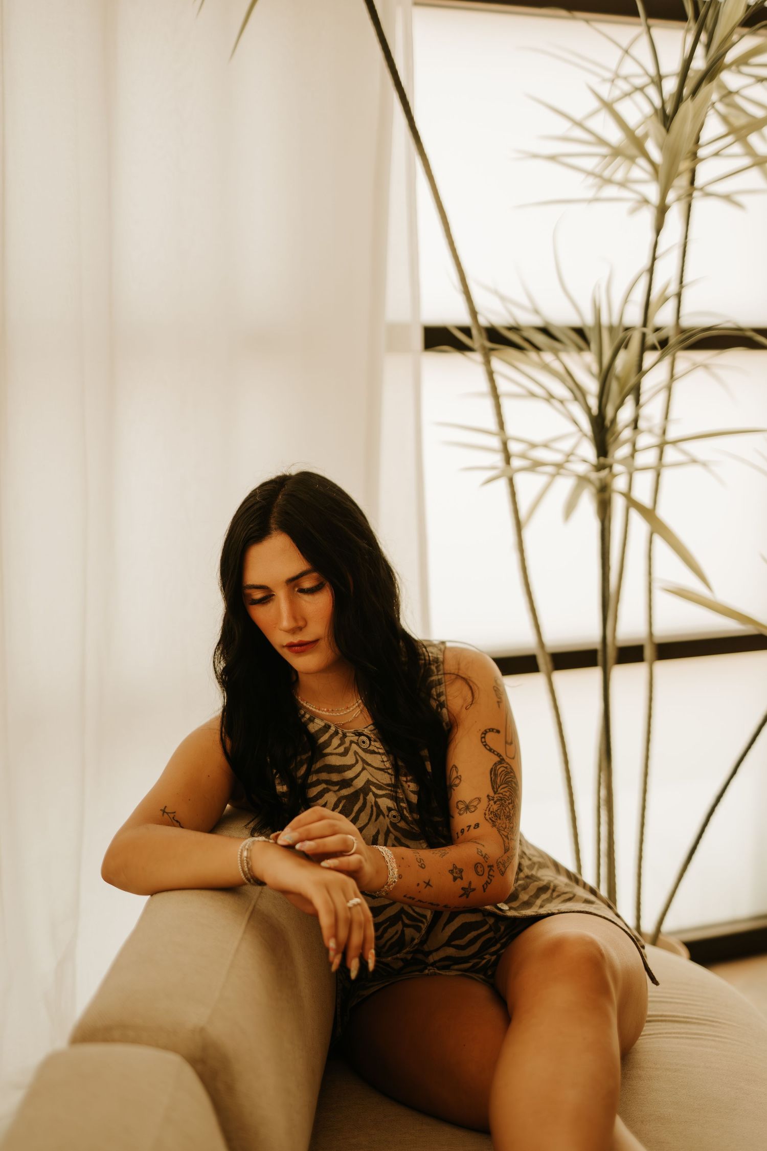 Tattooed woman in tiger print dress sits on sofa beside tall plant near bright window, looking down thoughtfully.