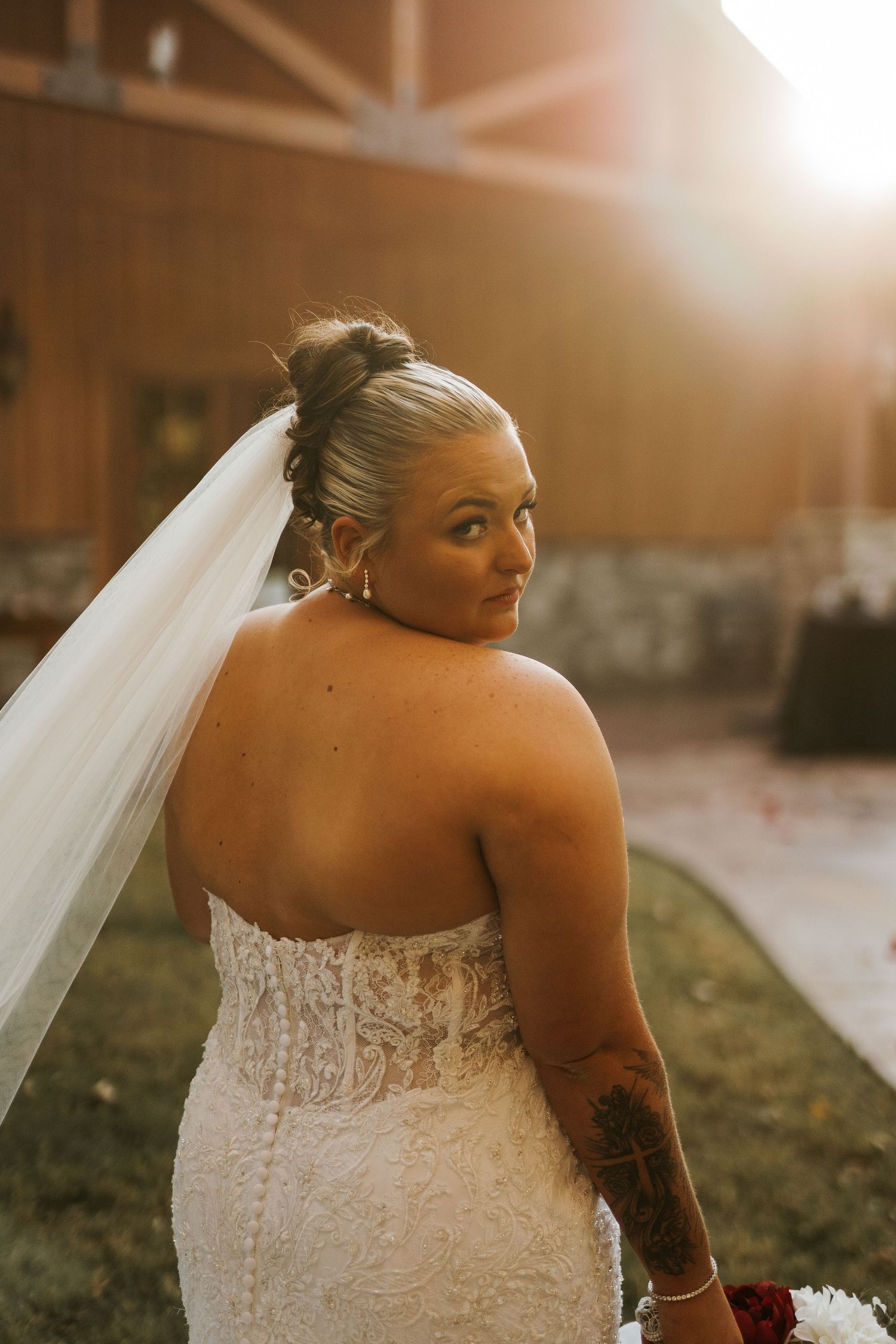 Bride in lace strapless gown and veil glances over shoulder in golden sunset light outdoors.